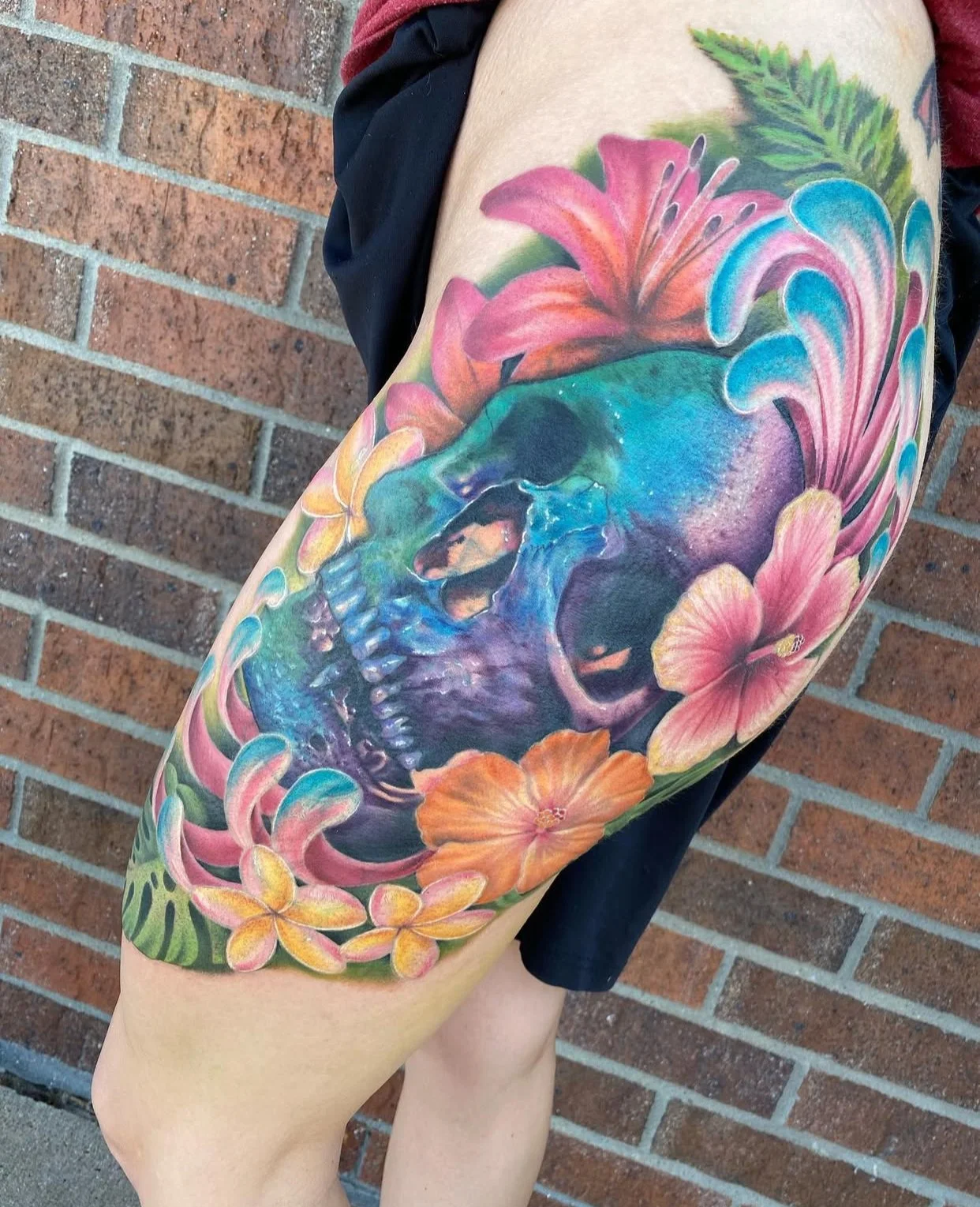 Colorful tattoo of a blue skull surrounded by pink, orange, and yellow flowers on a person's thigh, with a brick wall in the background.