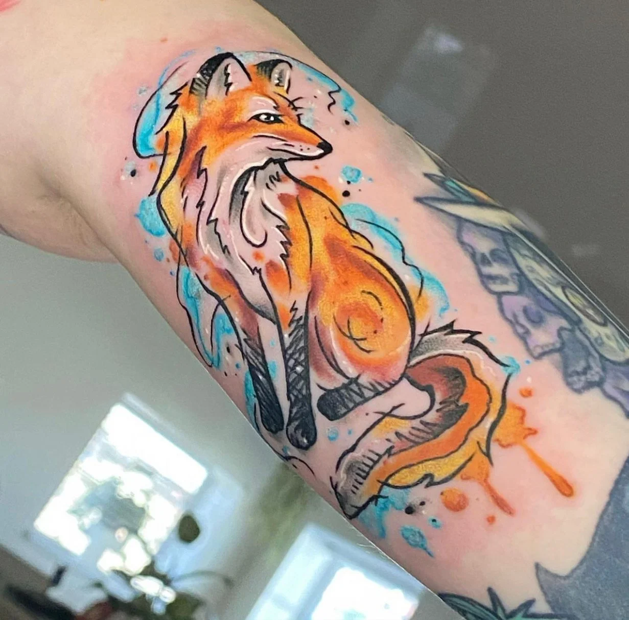 Colorful tattoo of a fox on a person's arm, with watercolor-style splashes of blue, orange, and black ink.