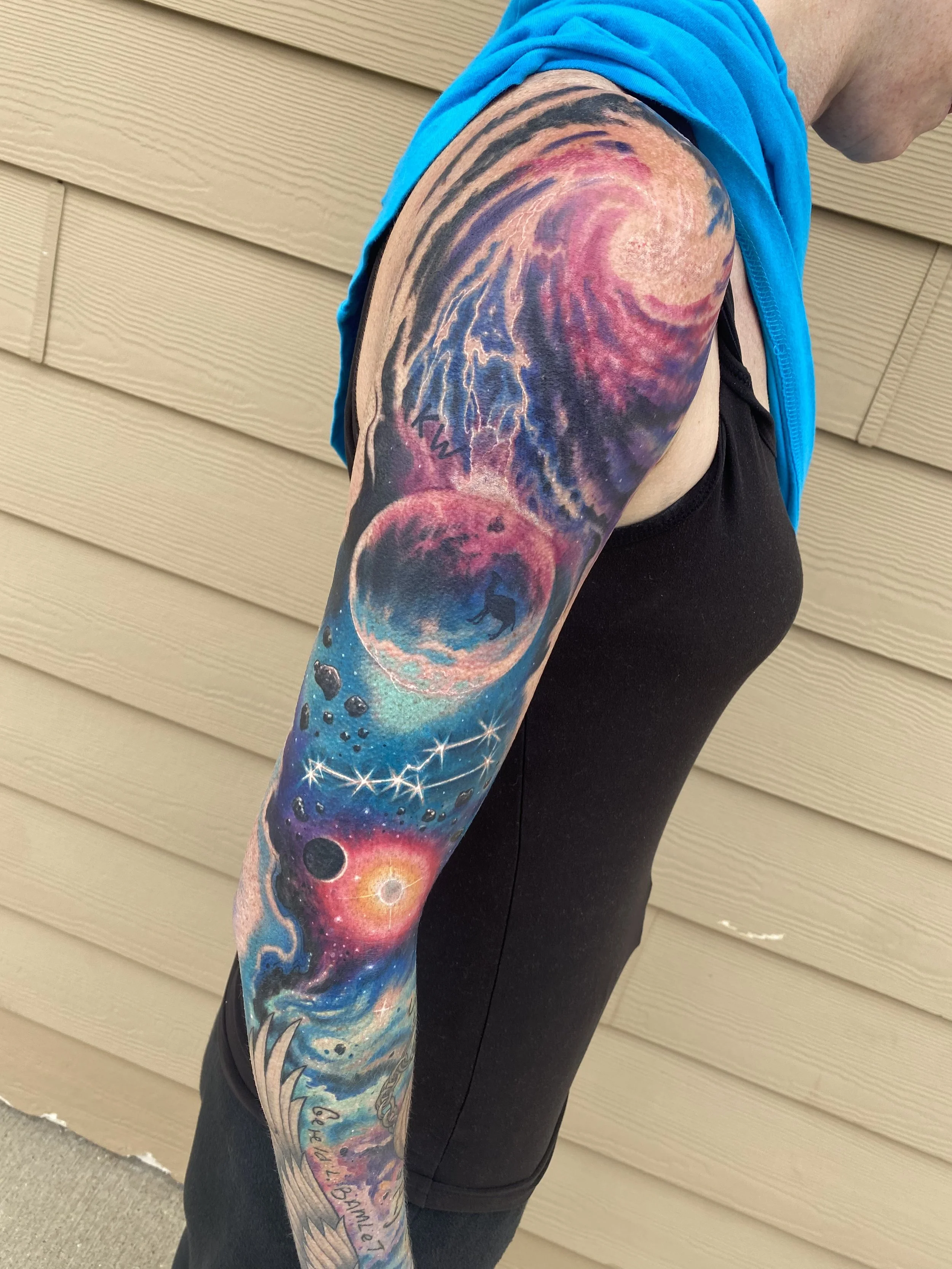 Colorful space-themed tattoo sleeve featuring a large planet, a wolf silhouette, stars, a solar eclipse, and swirling nebulae on a person's arm.