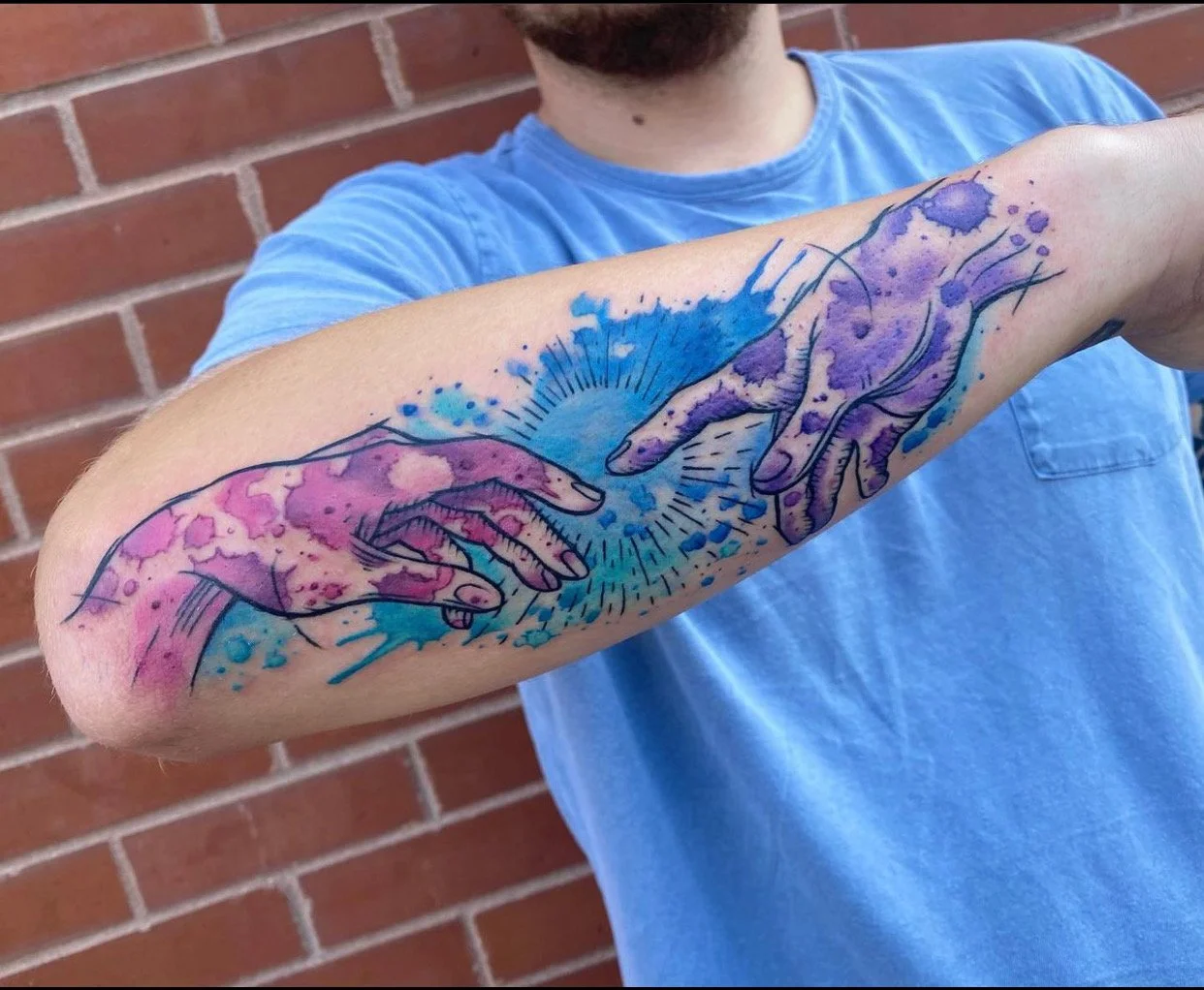 Tattoo of two hands reaching towards each other, with one in pink and the other in purple, surrounded by blue splashes of color.