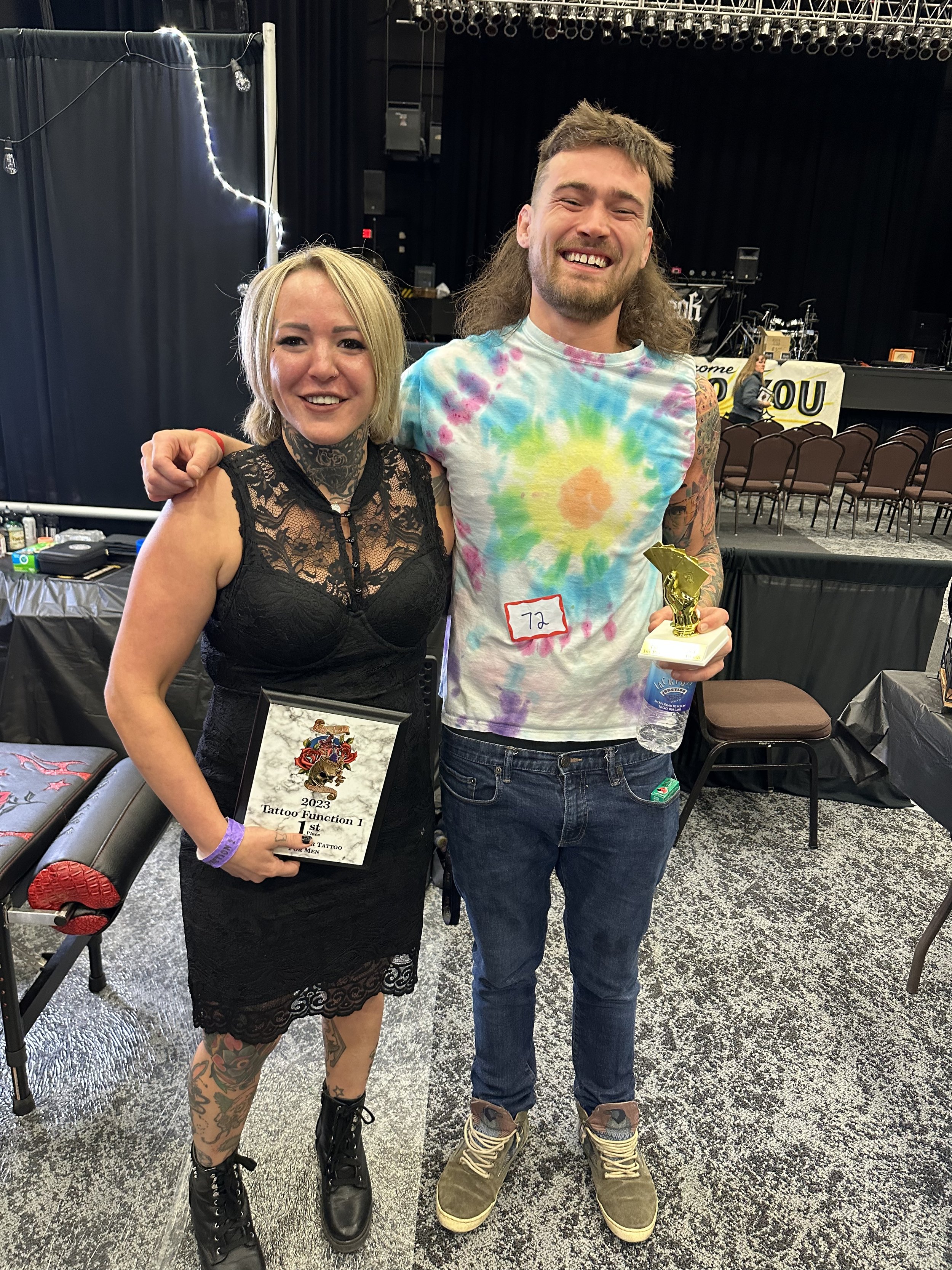 A woman and a man standing side by side at a tattoo event, smiling at the camera. The woman is holding a framed certificate and has tattoos and tattoos on her leg. The man is holding a small trophy and a water bottle, wearing a colorful tie-dye shirt, jeans, and sneakers.