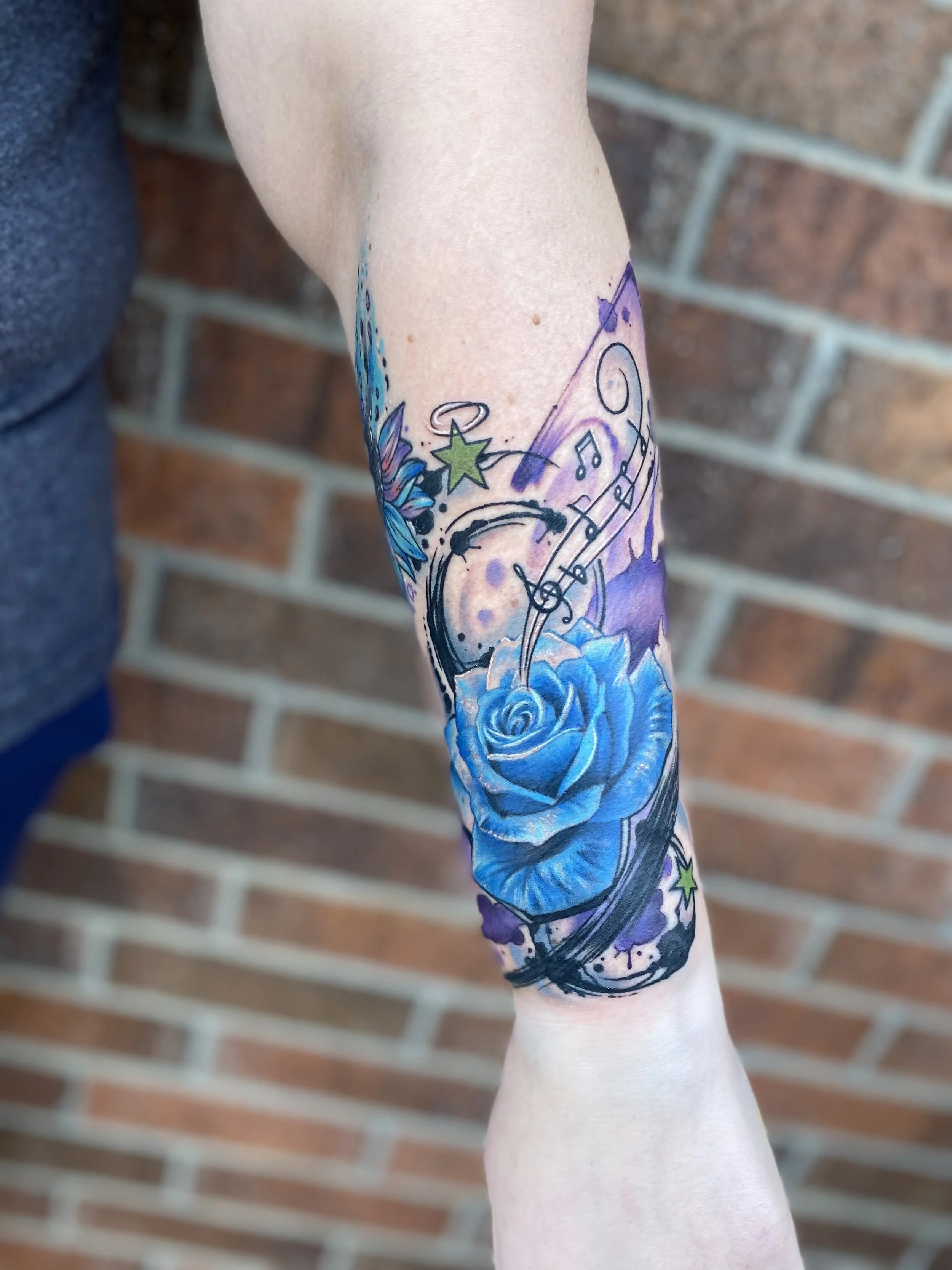 A colorful tattoo on a person's forearm featuring a blue rose, musical notes, stars, and abstract splashes of color against a brick wall background.