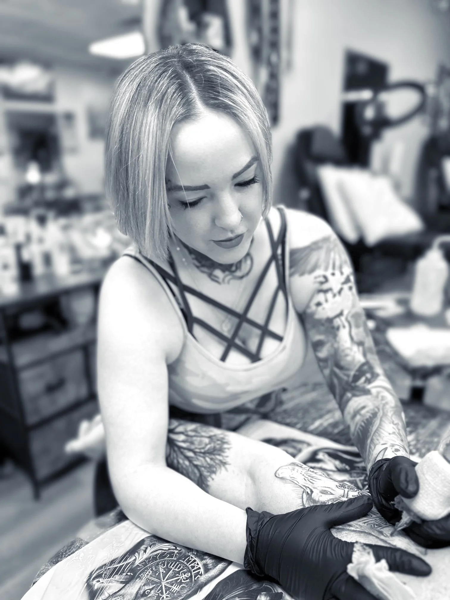 A woman with tattoos and blonde hair is sitting at a table, wearing black gloves, and getting a tattoo on her arm.