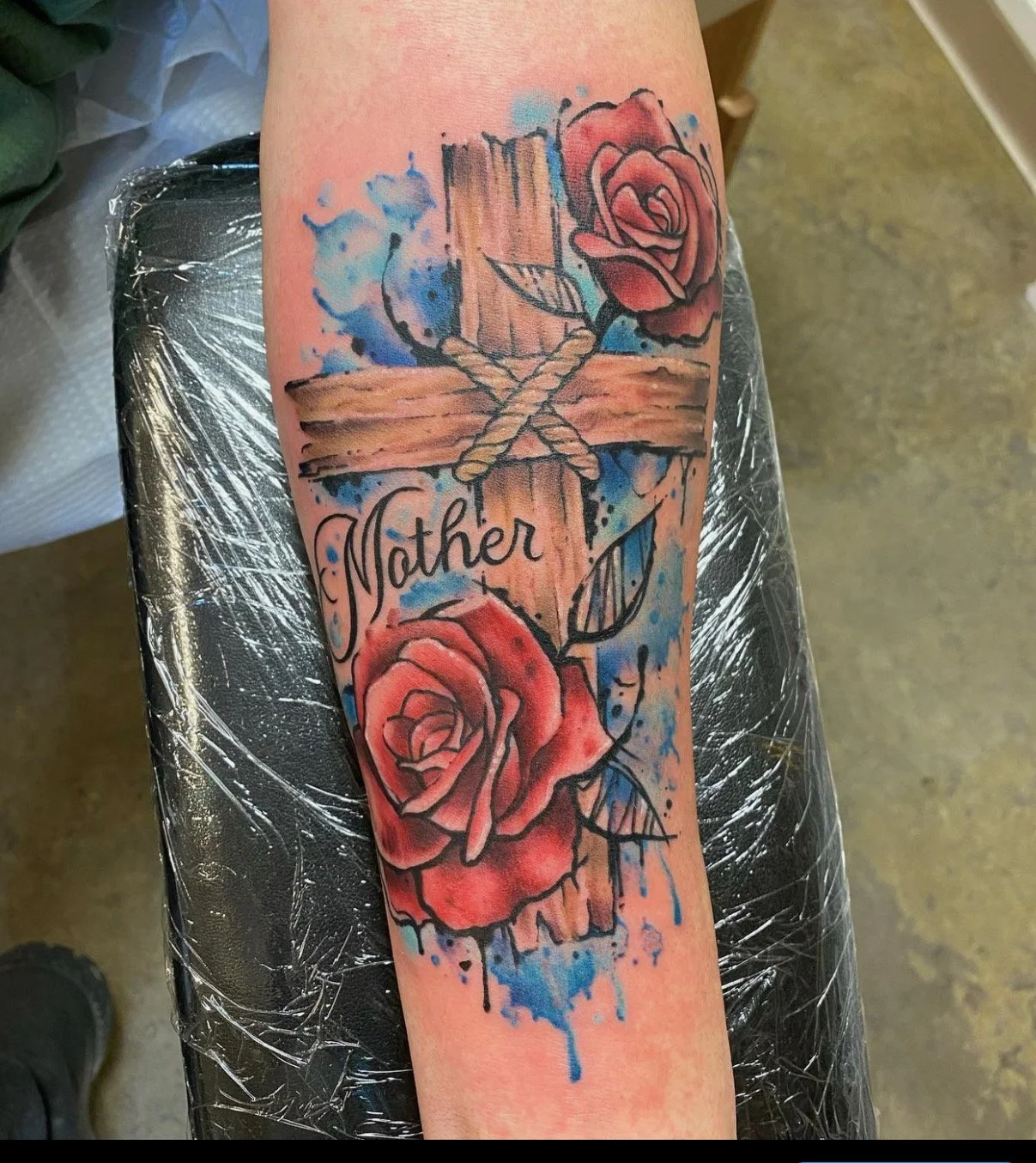 A tattoo of a wooden cross with roses and the word 'Mother' on a person's arm.