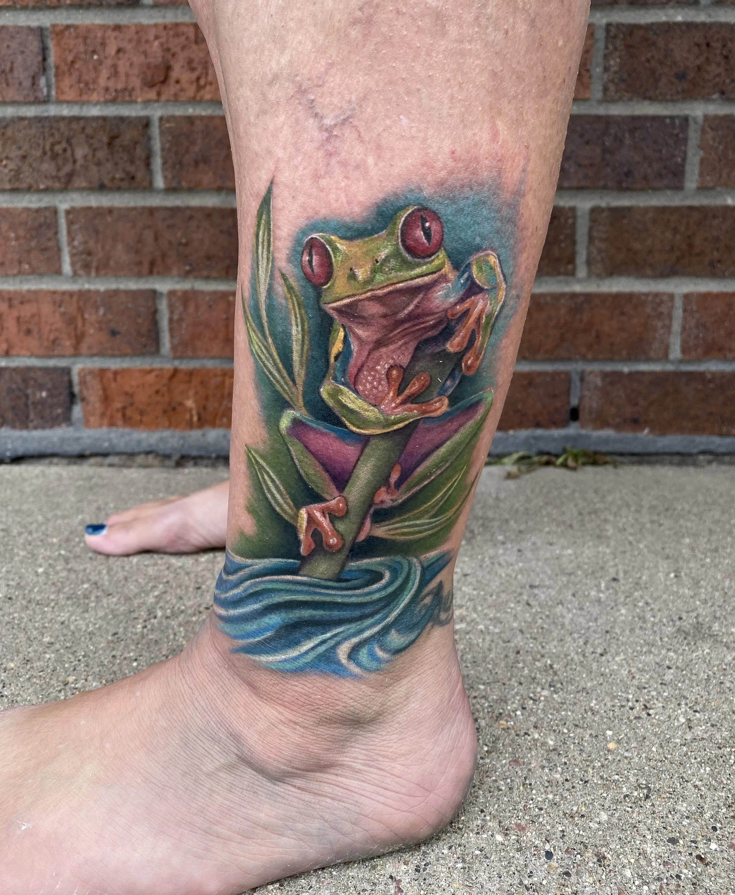 Colorful tattoo of a frog sitting on a plant stem above water, with the frog's large red eyes and open mouth visible.