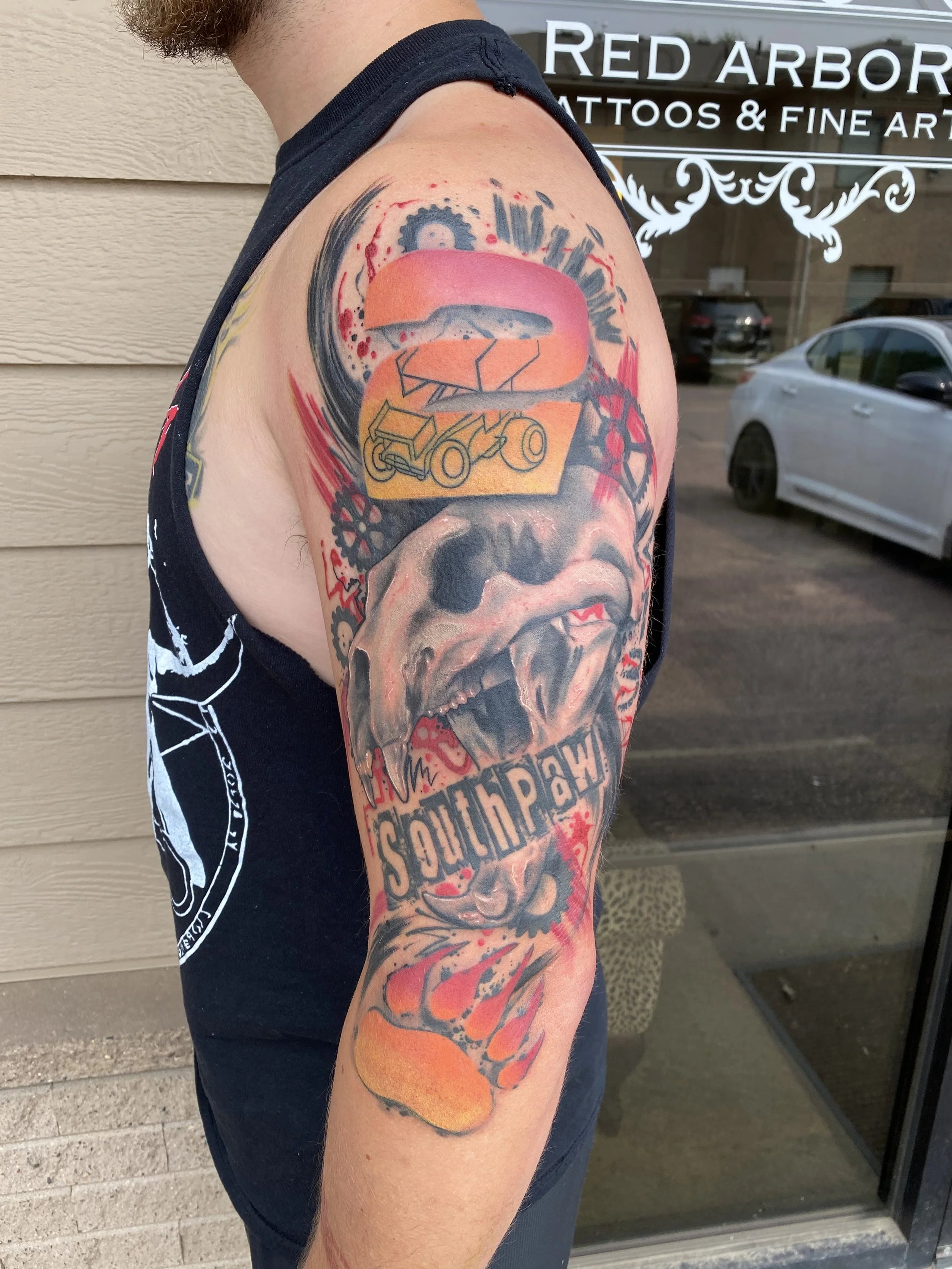 Tattoo of a snarling dog with the word "Southpaw" on the upper arm, decorated with flames and red accents, and a colorful abstract design with gears and a vehicle above the dog.