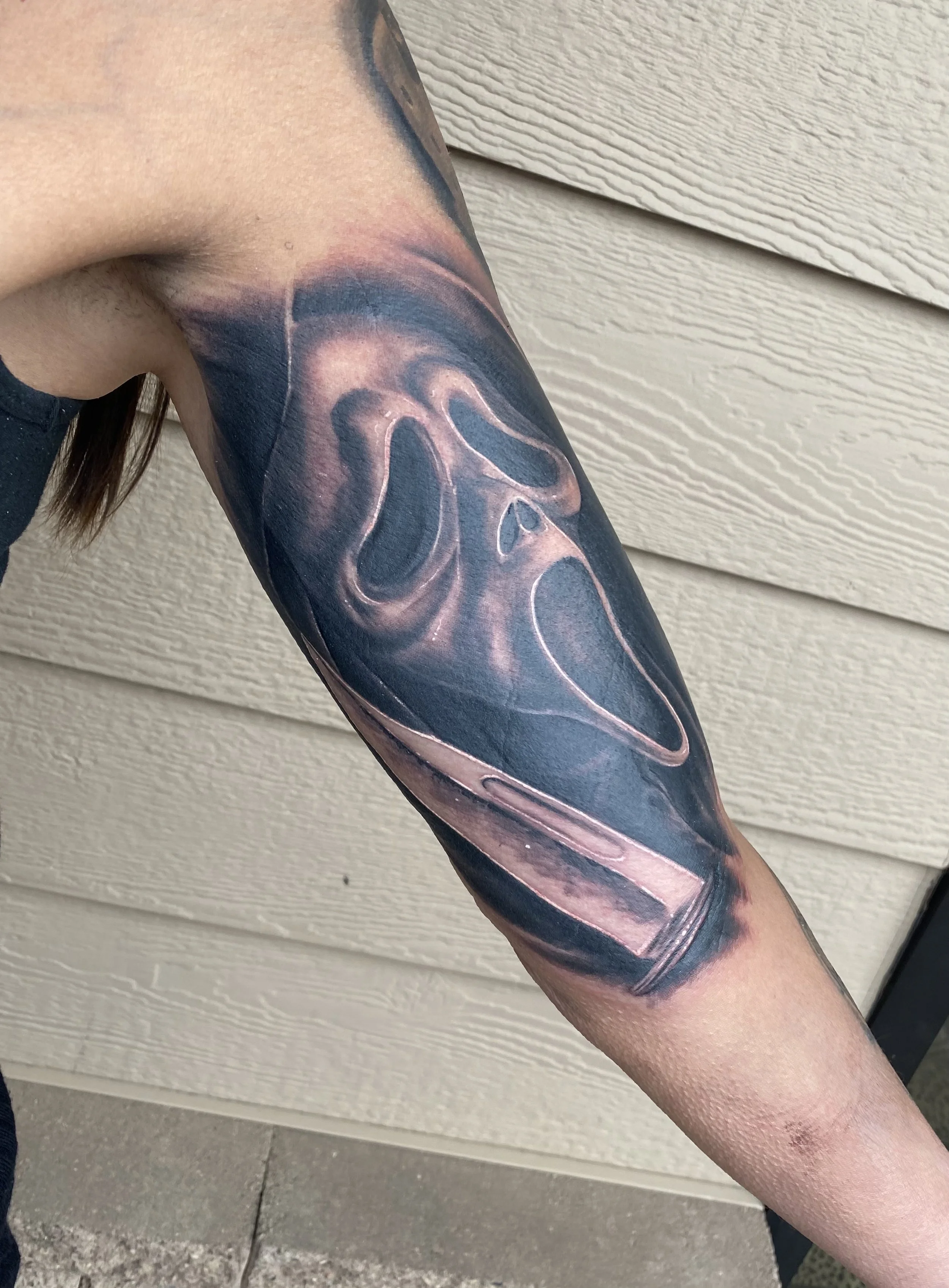 A tattoo of the iconic ghost face mask from the Scream movie franchise, with a background of a black and pinkish hue, covering the person's upper arm.