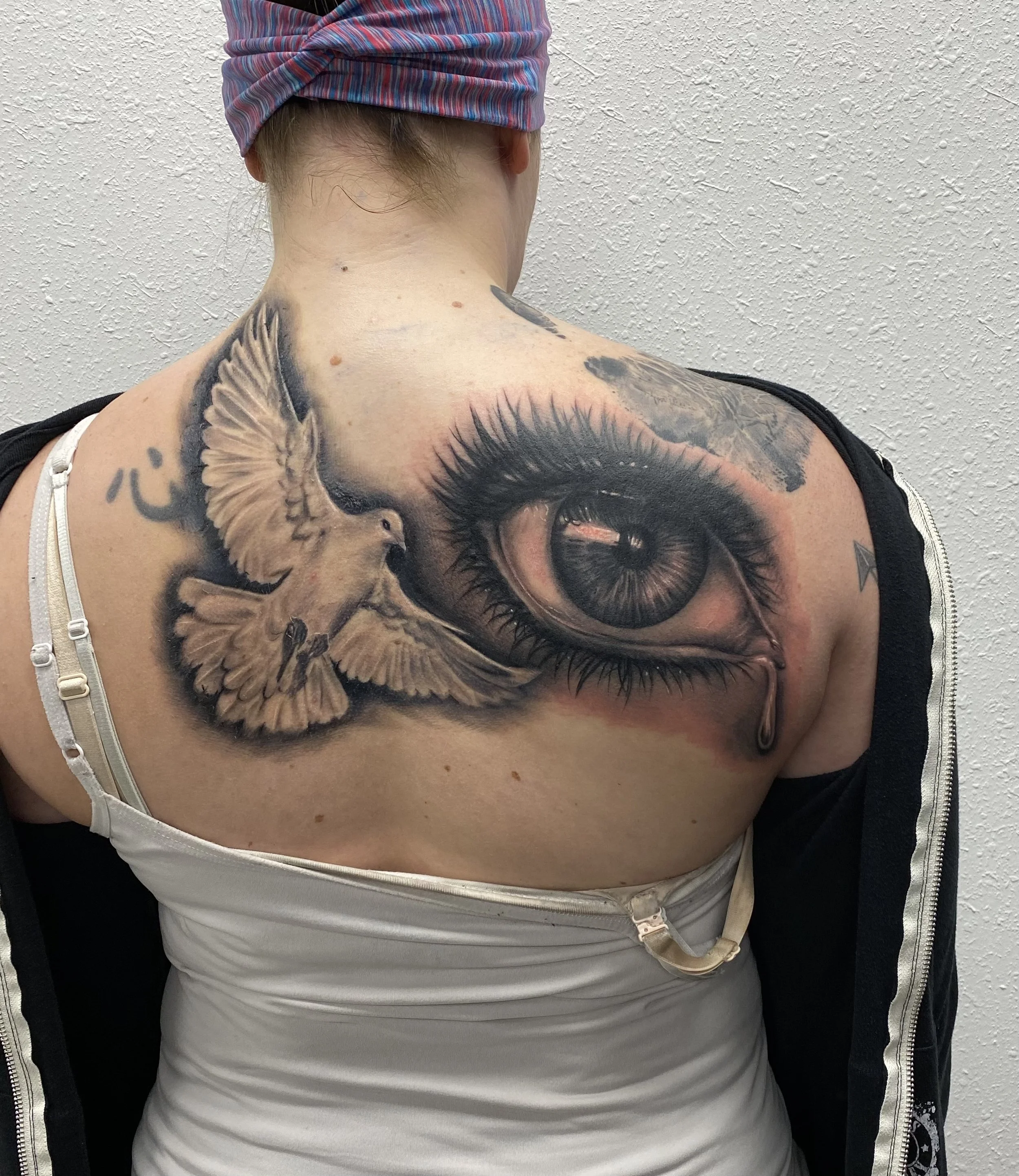 Tattoo of a realistic eye with tears and a flying dove with outstretched wings on a person's shoulder and upper chest.