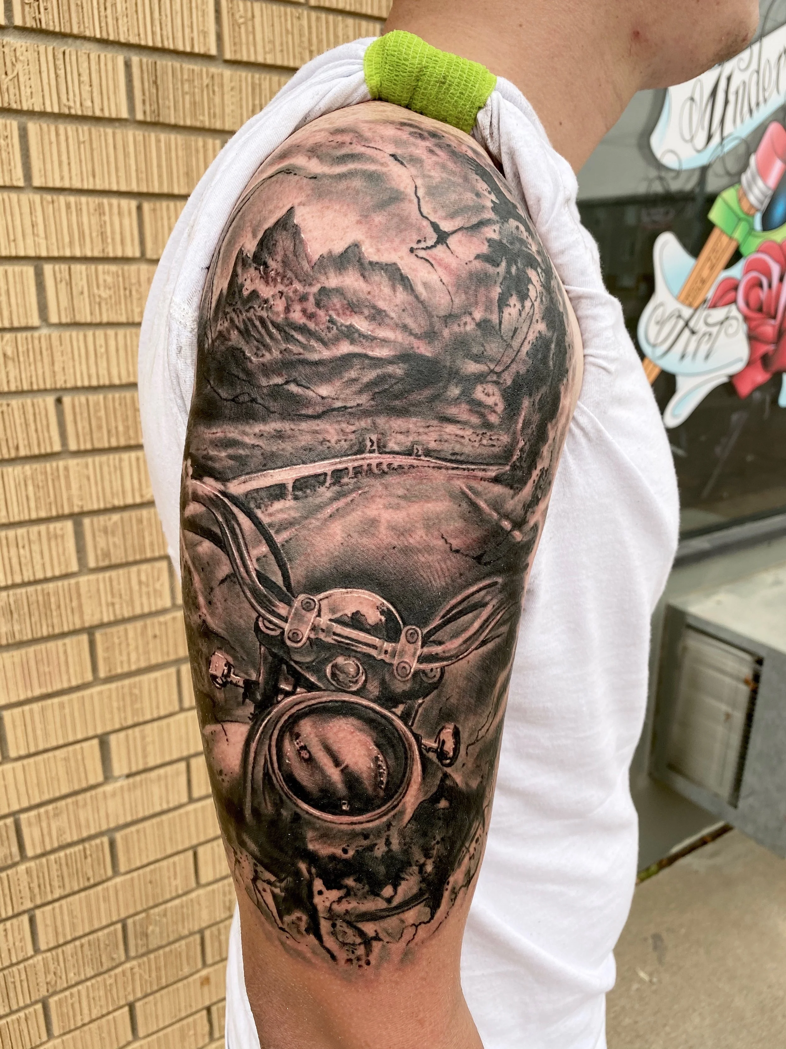 Black and gray tattoo of a motorcycle on a person's upper arm, with a landscape and cloudy sky background.