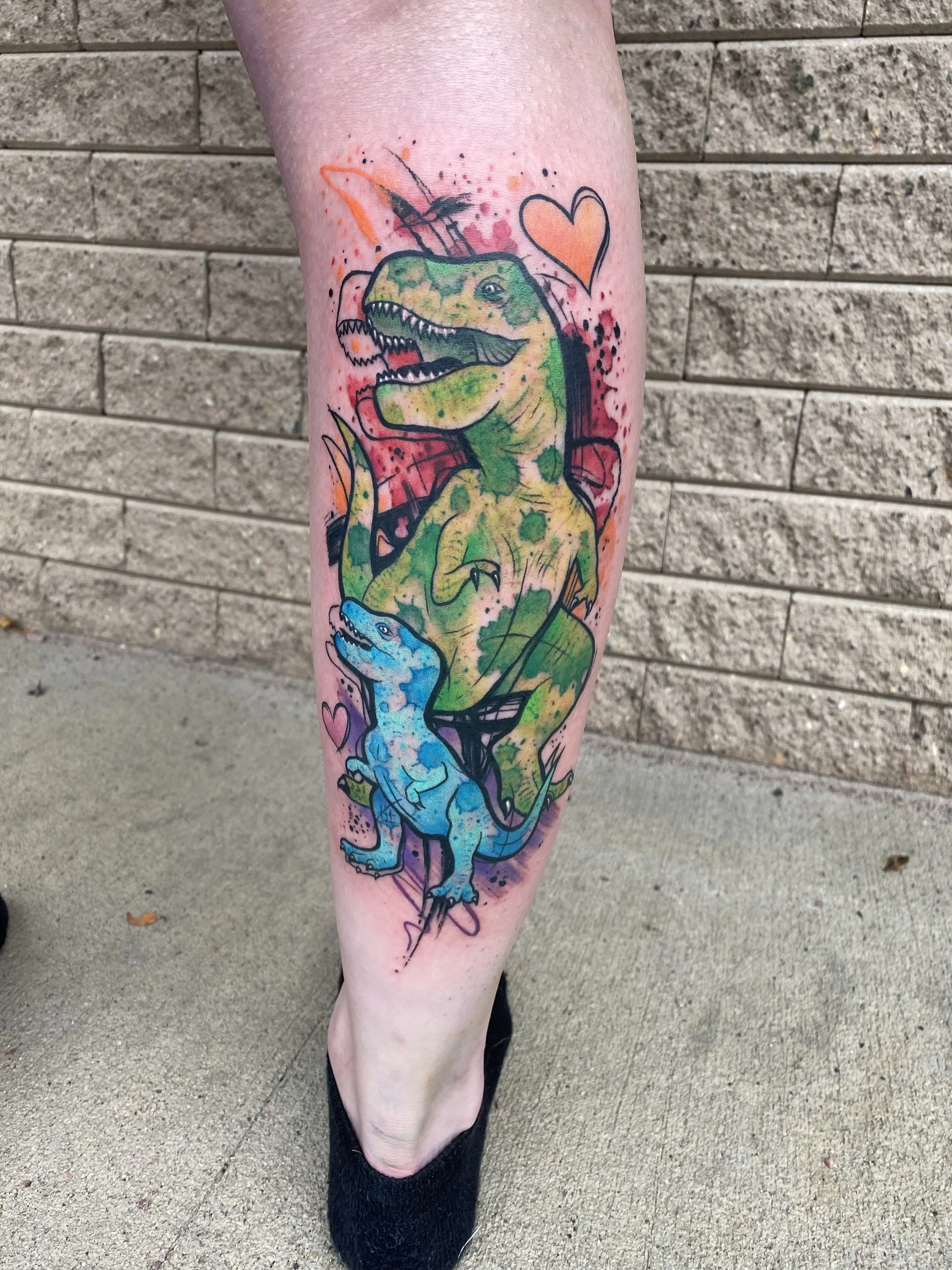 Tattoo of a green dinosaur with a smaller blue dinosaur, both with detailed features, surrounded by hearts and colorful splashes on a person's calf.
