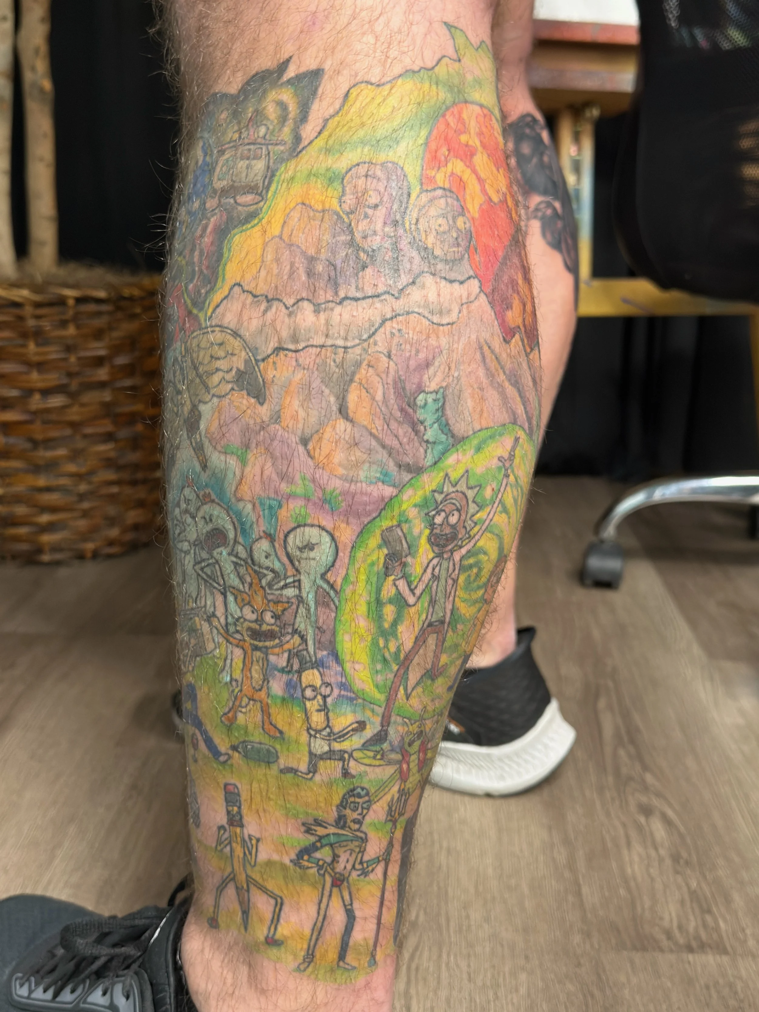Sioux Falls South Dakota Tattoo Shop. Apex Ink. Colorful tattoo on a person's calf depicting various characters from animated shows and movies, including Rick and Morty, aliens, and a rainbow.