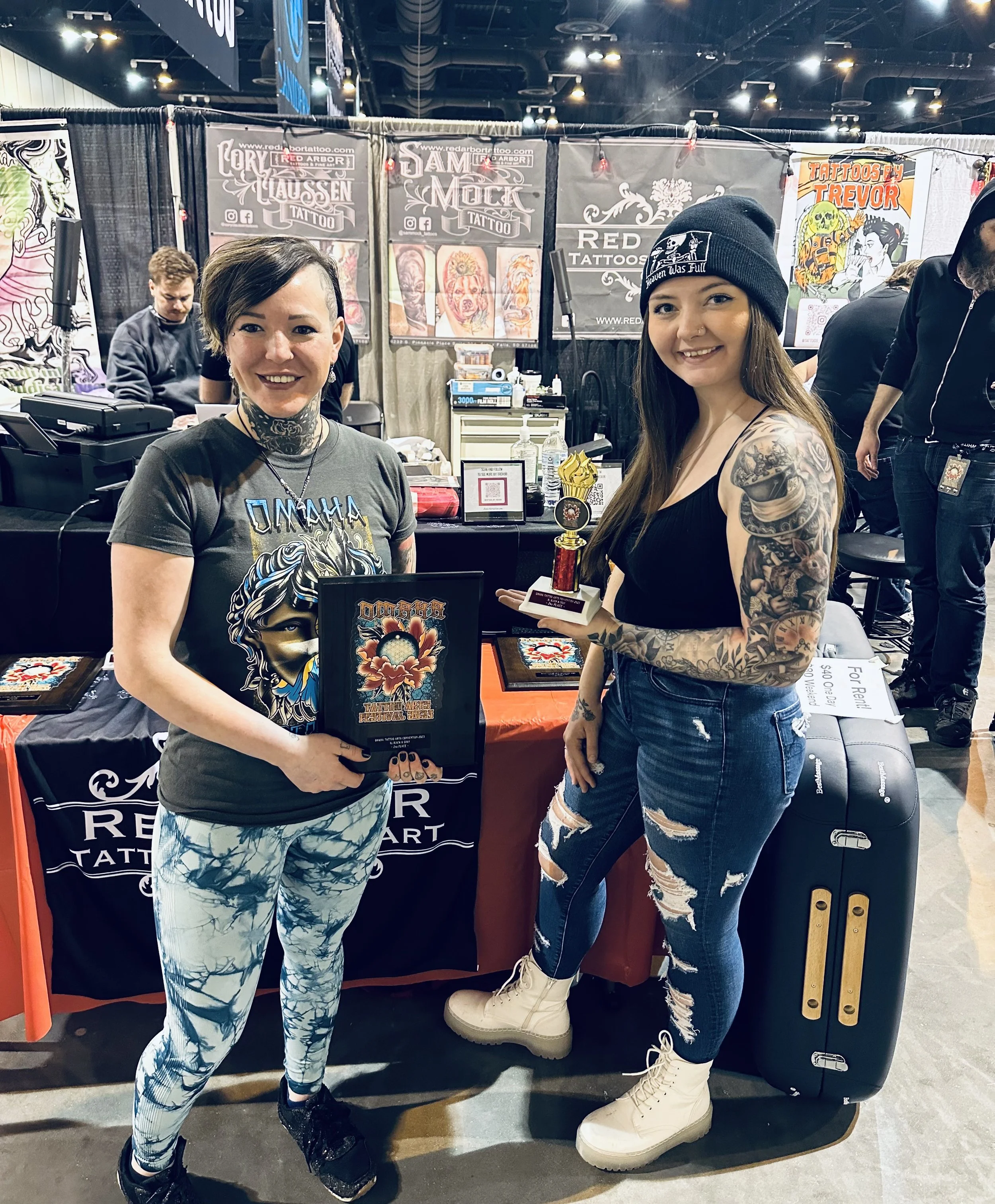 Two women with tattoos at a tattoo convention, one holding a trophy. They are standing in front of a booth featuring tattoo art and banners.