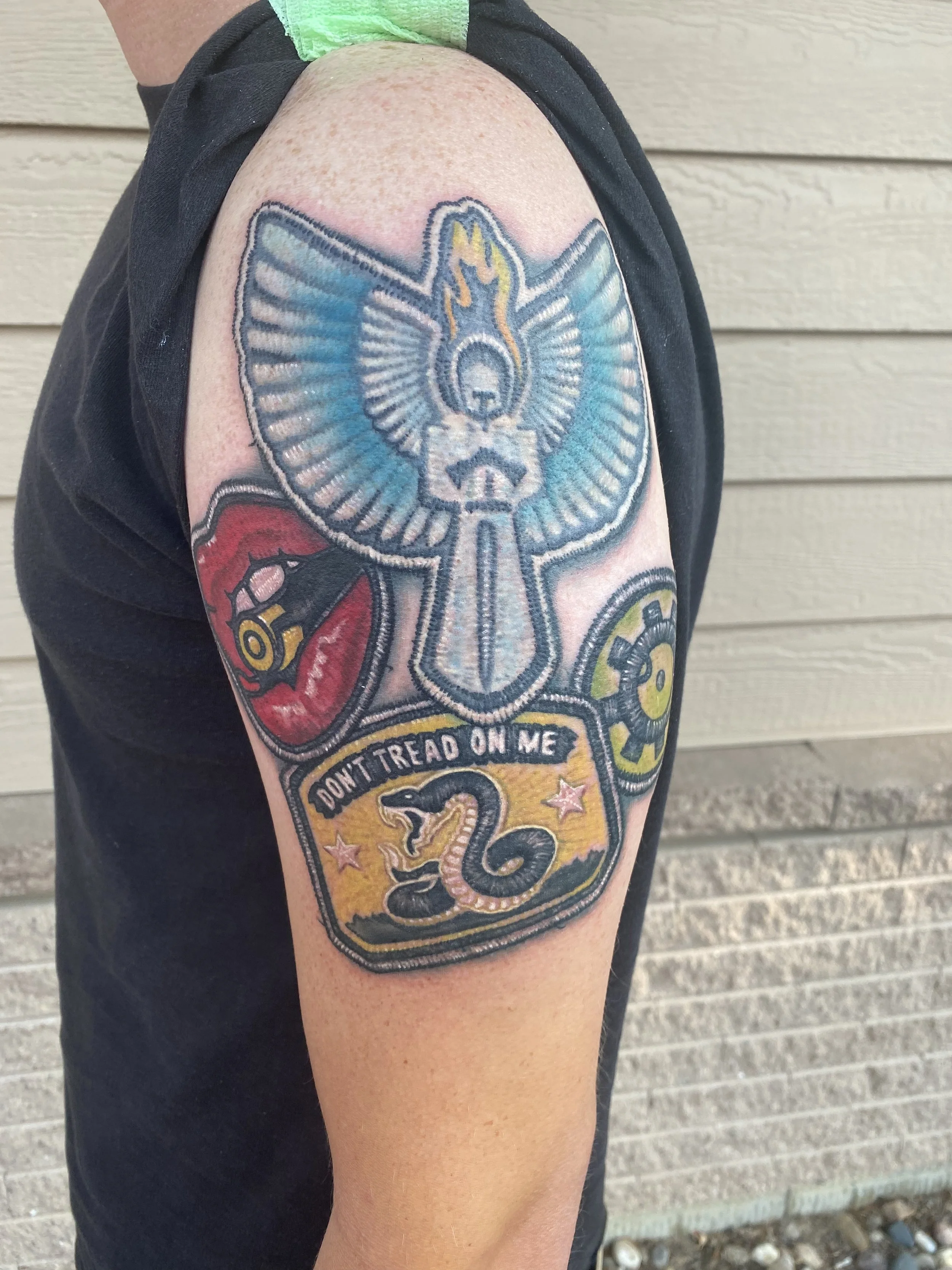 Colorful tattoo on upper arm featuring a large angel with outspread wings and a flaming sword, a snake with the phrase 'Don't Tread on Me', a skull, a gear, and a red heart with a shattered emblem.