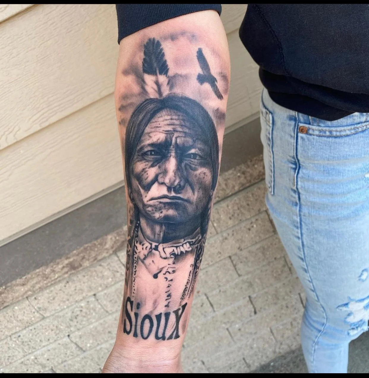 Tattoo of a Native American man with long hair, feathers, and traditional clothing, with the word "SIOUX" at the bottom.