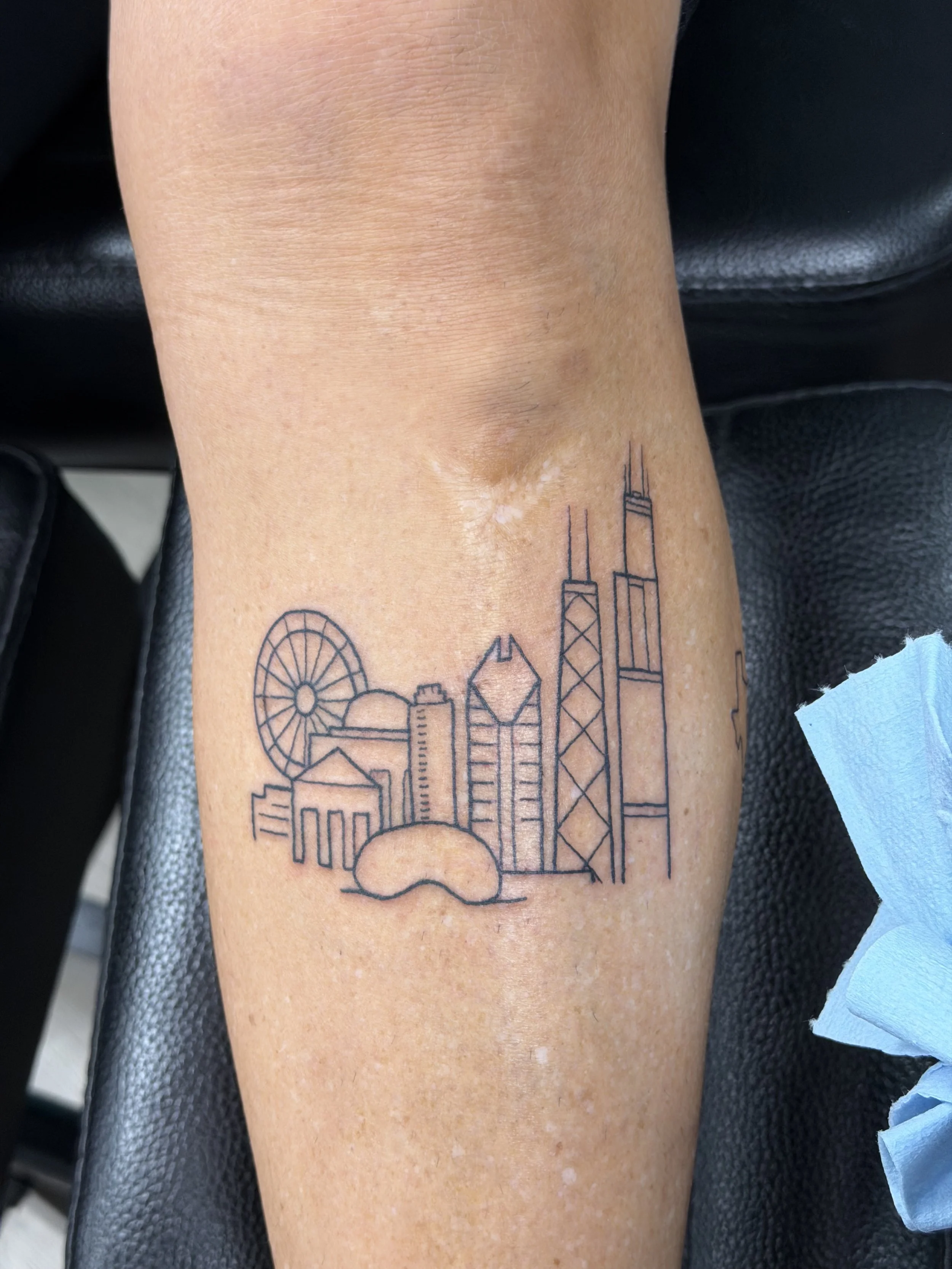 Tattoo of a minimalistic city skyline in black ink on a person's arm, featuring buildings, a Ferris wheel, and a bridge.