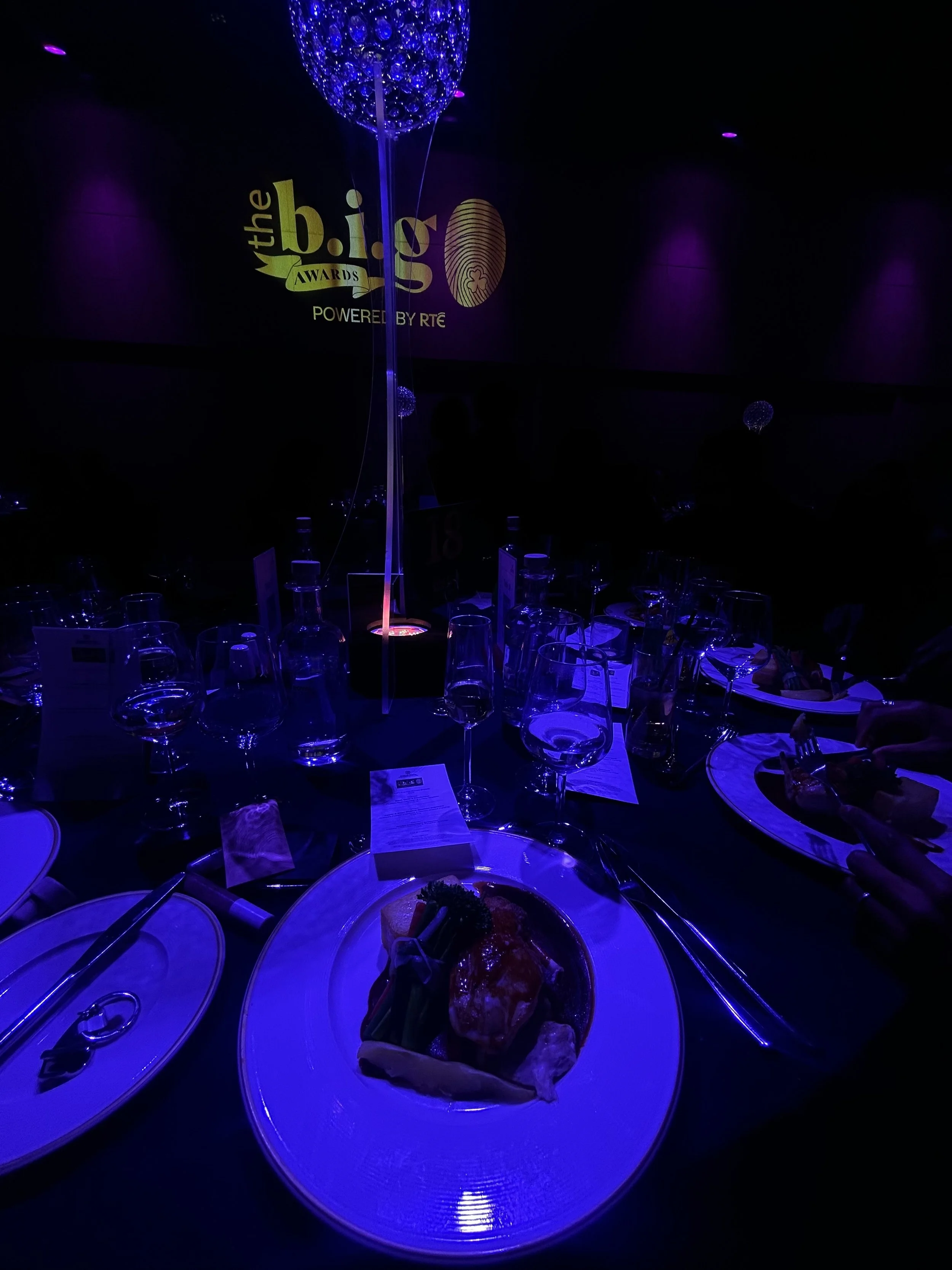 A dimly lit banquet table at an awards event with a purple theme. The table has wine glasses, plates, and a centerpiece with a helium balloon. A screen in the background displays the logo for 'the b.i.g Awards' powered by RTE.