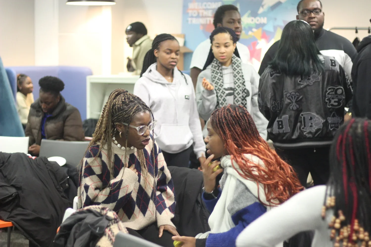 A group of diverse young women and girls are engaged in conversation in a crowded indoor setting. Two women are sitting in the foreground, talking intently, while others stand around them, some looking at their phones or listening.