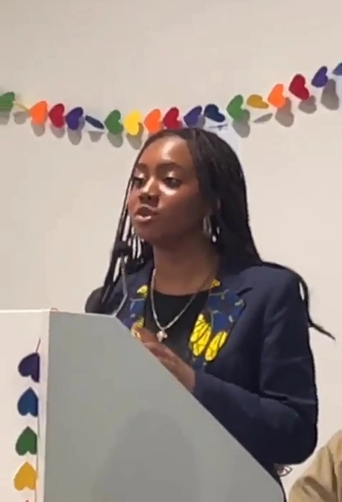 A woman with long braided hair standing behind a white podium with colorful heart-shaped decorations, speaking at an event.