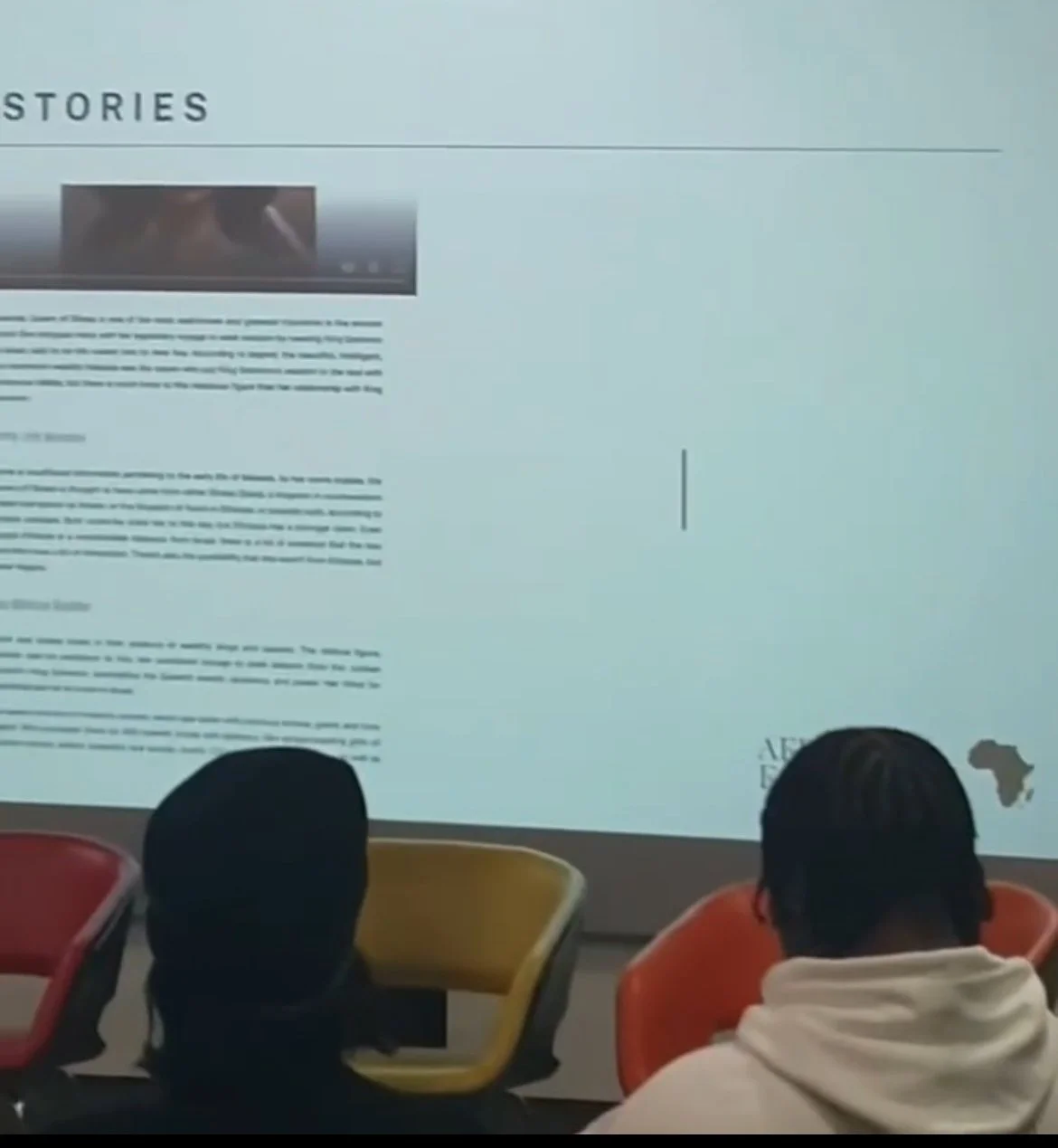 Two people sitting in front of a large screen display, which shows a webpage titled 'STORIES' with text and a small map of Africa.