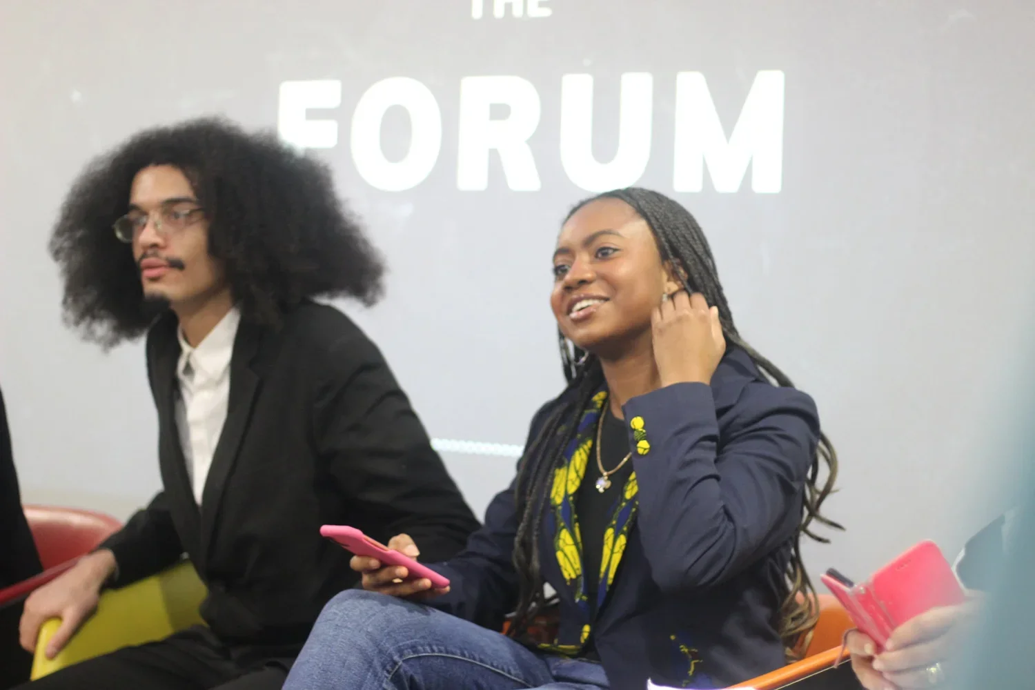 Two people sitting in a conference room with a gray wall behind them. A man with long curly hair, glasses, and a suit is on the left, and a woman with long braided hair, wearing a dark jacket and holding a pink phone, is on the right. Part of the tex