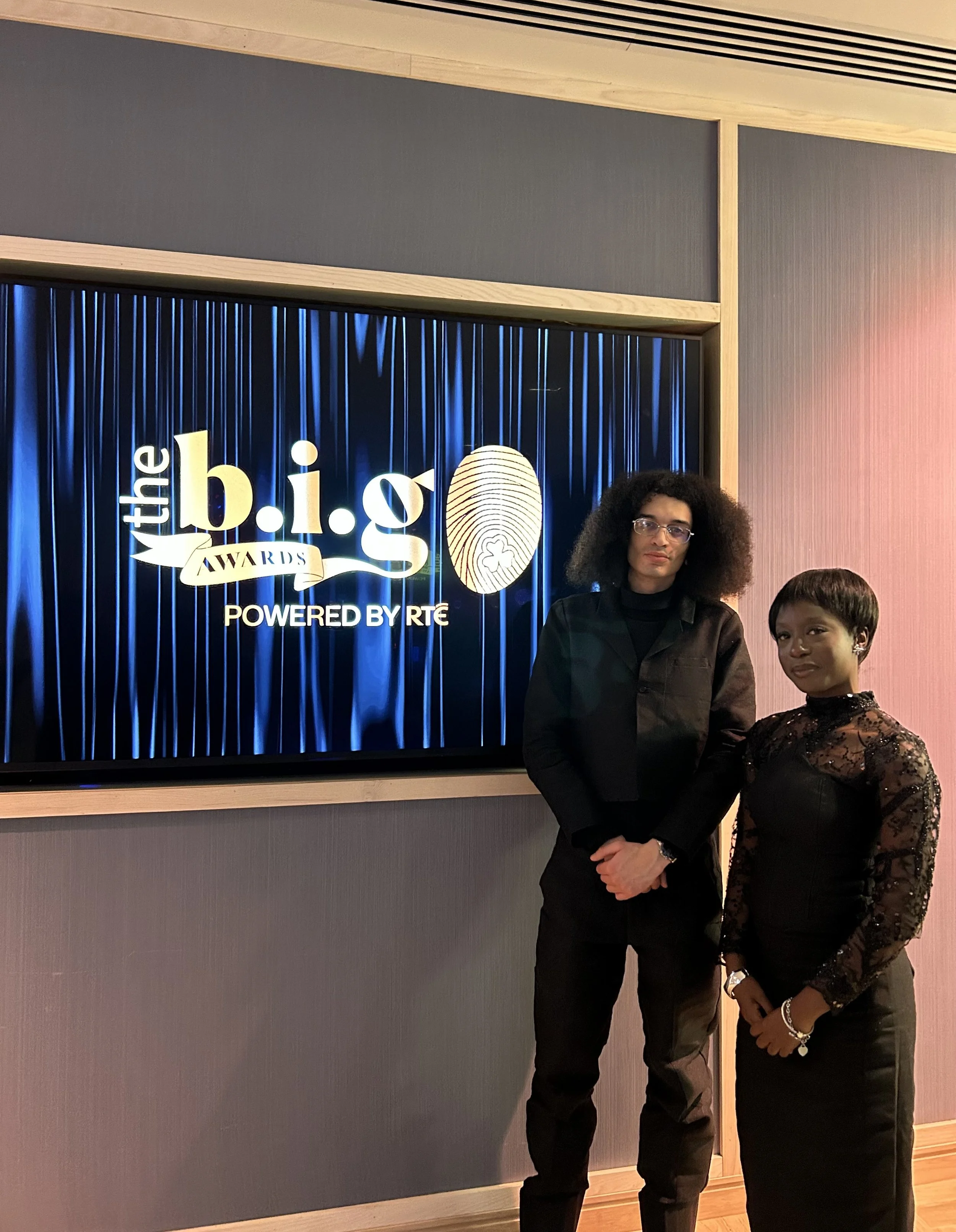 Two women standing in front of a sign for the b.i.g awards, powered by RTE. The sign has a blue curtain background and a fingerprint logo.