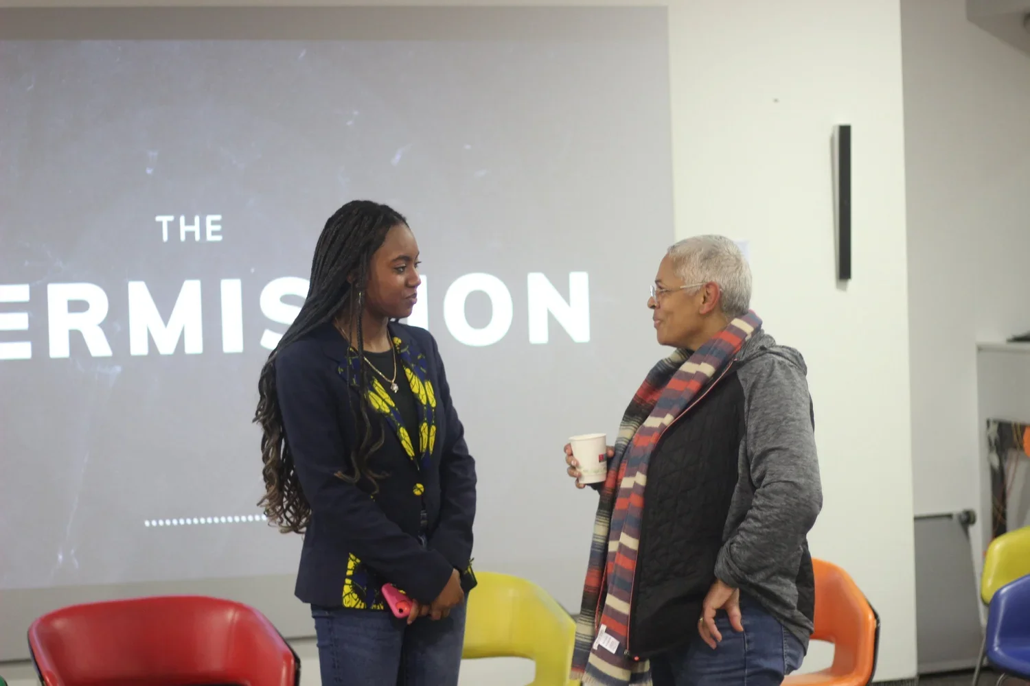 Two women standing and talking in a room with a presentation screen that says 'The PermissioN'. One woman, younger with long braided hair, dressed in a navy blue jacket with yellow patterns, holding a pink object. The other woman, older with short gr