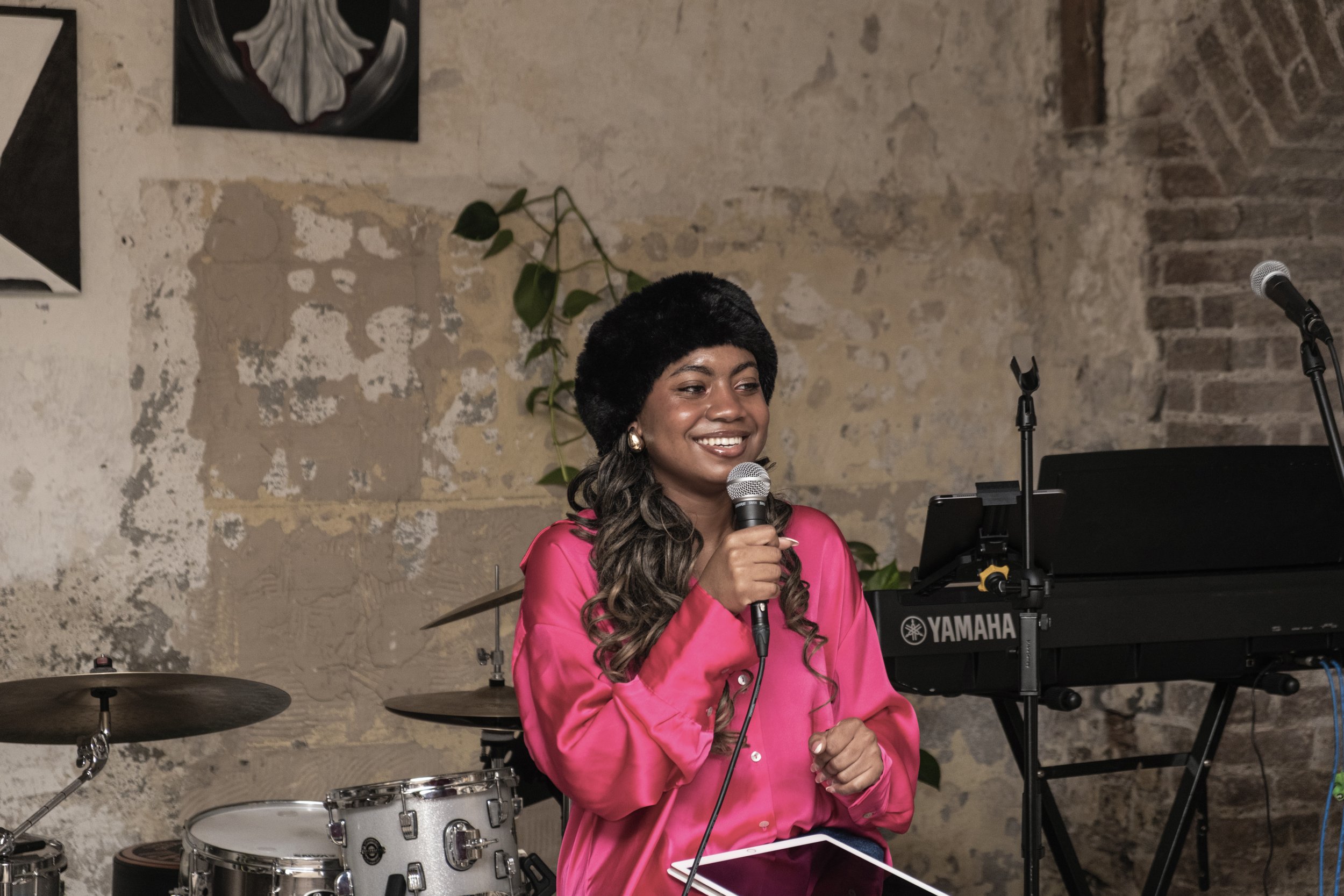 A woman smiling and holding a microphone while sitting near a keyboard and drums in a rustic setting.