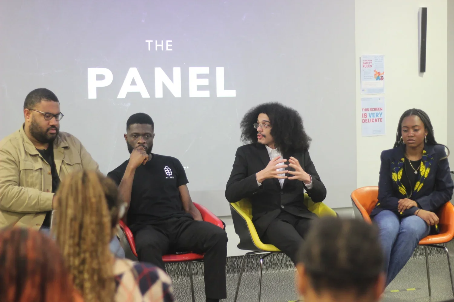Panel discussion with four diverse individuals seated on stage in a room with a gray wall background and a screen displaying 'The Panel.'
