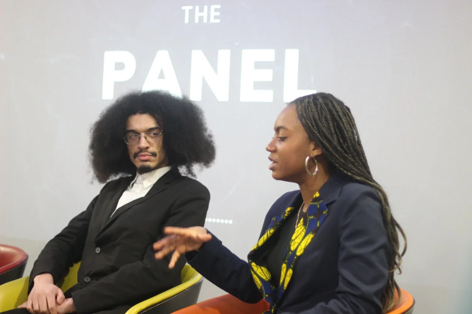 A man and a woman are sitting and talking during a panel discussion, with a projection behind them that reads 'The Panel'.