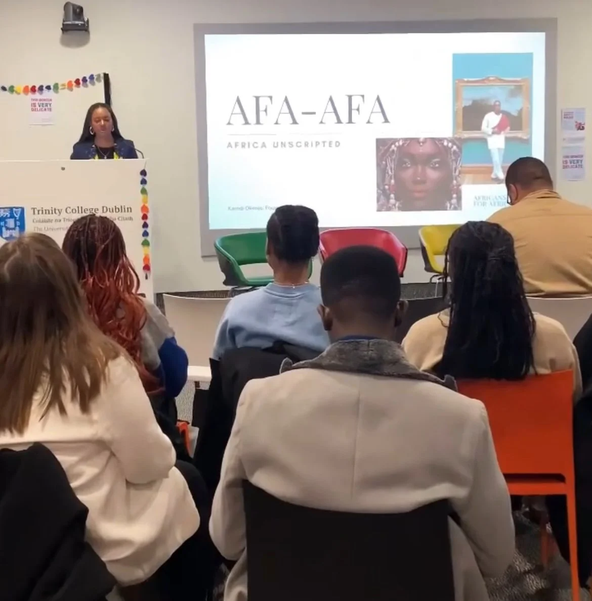 A woman presents at a lecture titled 'AFA-AFA' at Trinity College Dublin, with a screen displaying images of African art and culture, and an audience listening attentively.