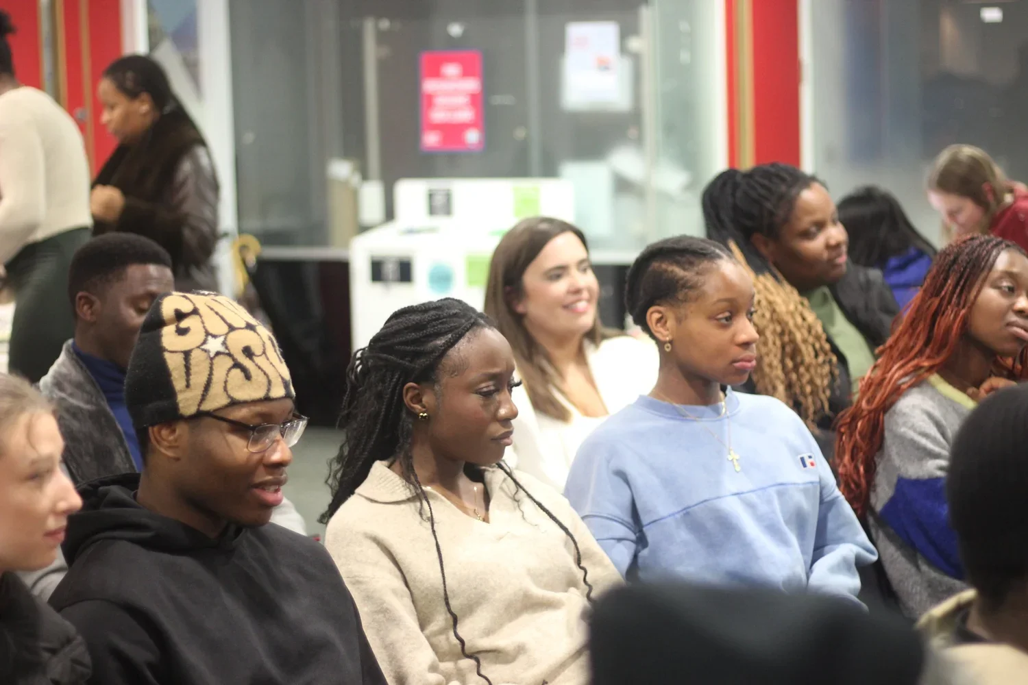 A group of diverse young women sitting in a room, attentively listening. Some are focused, others appear slightly distracted. They are dressed casually and have various hairstyles.