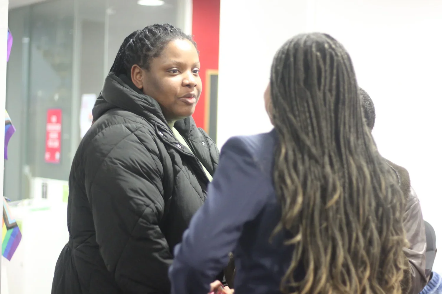 Two women chatting indoors, one facing the camera with her face visible and the other with her back turned, both wearing jackets.