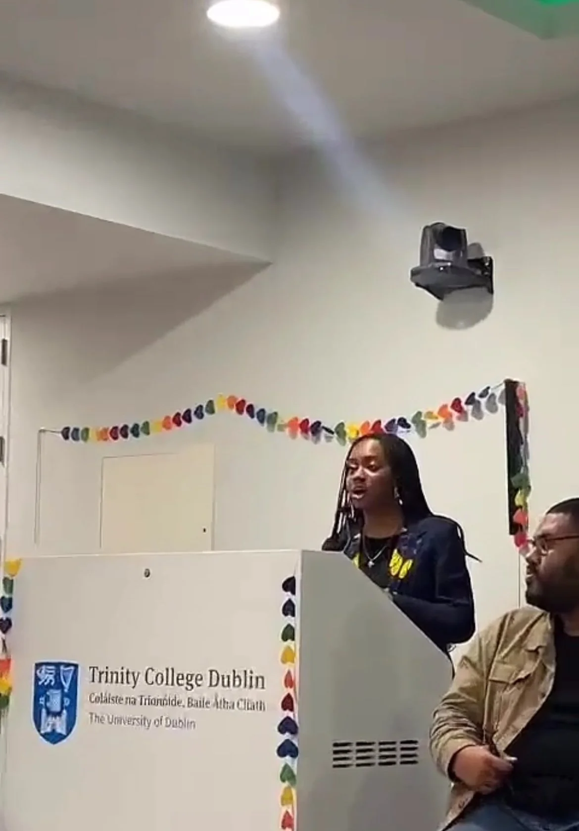 A woman speaking at a podium decorated with rainbow-colored hearts at Trinity College Dublin. A man is sitting next to her, listening. The podium displays the Trinity College Dublin logo and a banner with the college's name in Irish and English.