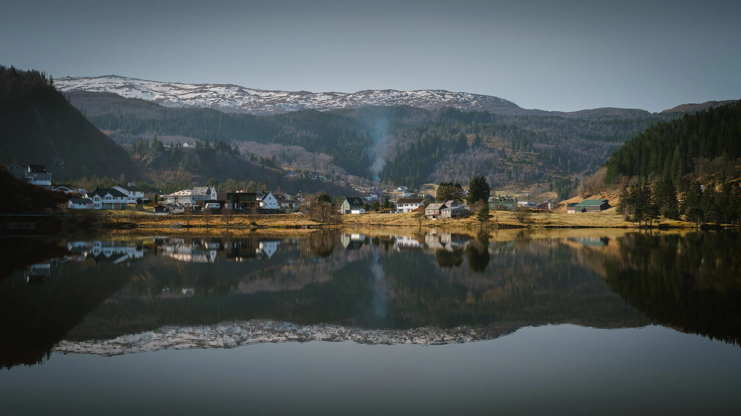 Paisagem tranquila na Noruega com água espelhada, pequena povoação e floresta entre montanhas.