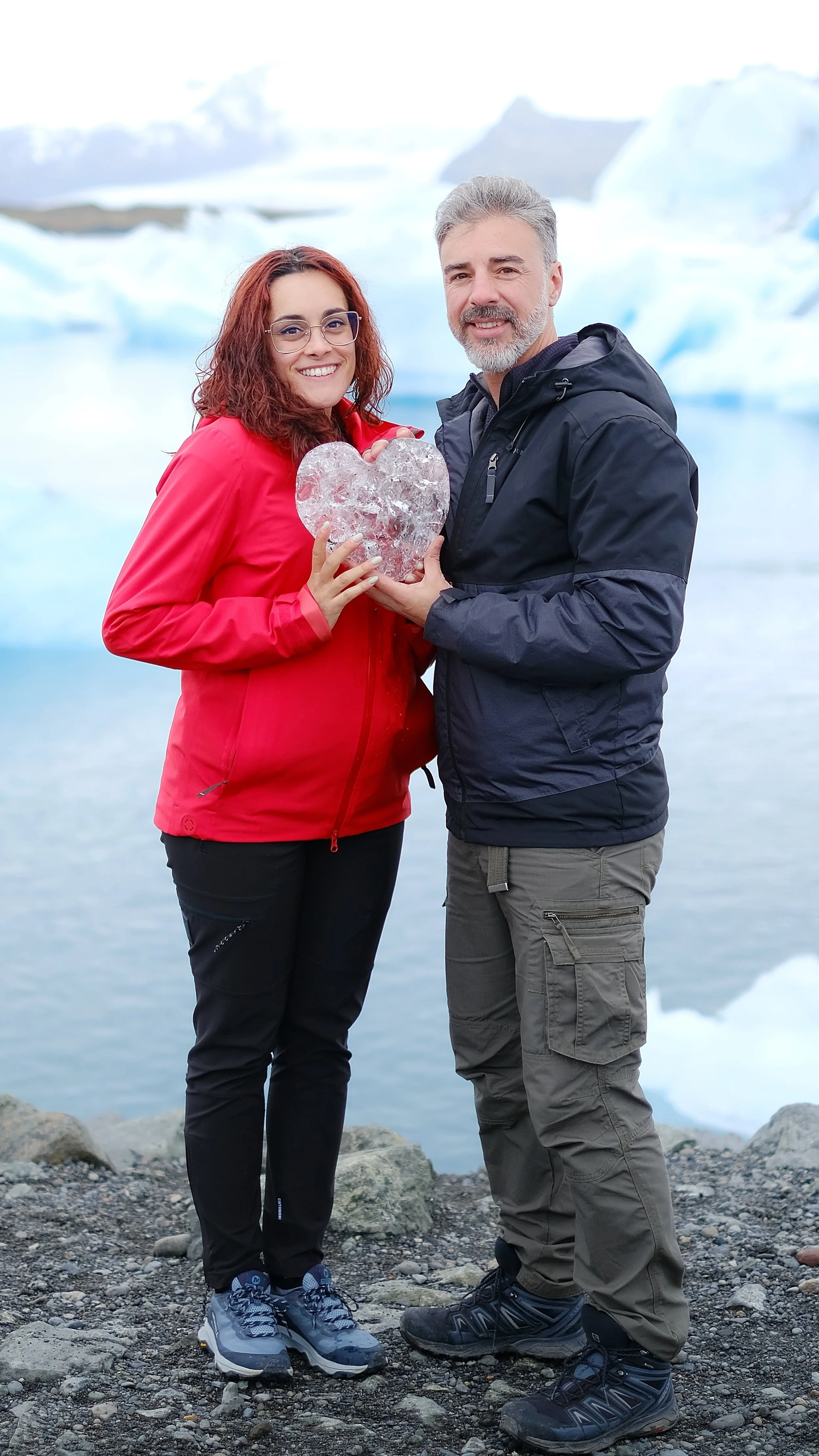 Casal junto à lagoa glaciar Jökulsárlón, na Islândia, a segurar um coração de gelo com icebergs ao fundo.