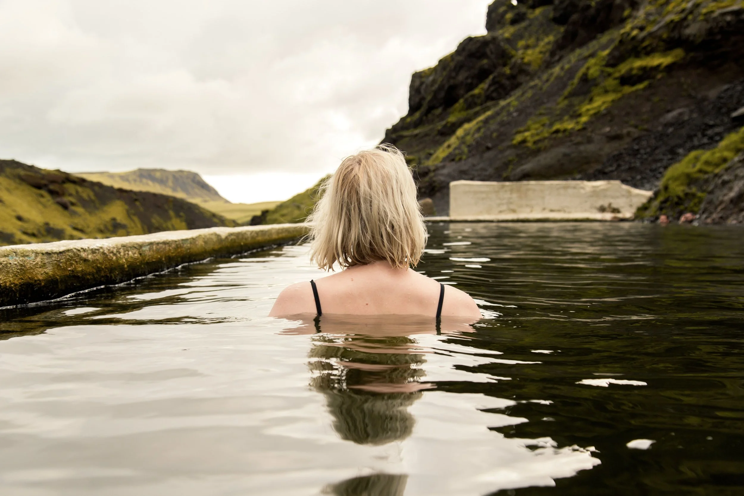 Mulher a relaxar numa piscina geotermal na Islândia, rodeada por montanhas e uma paisagem tranquila.