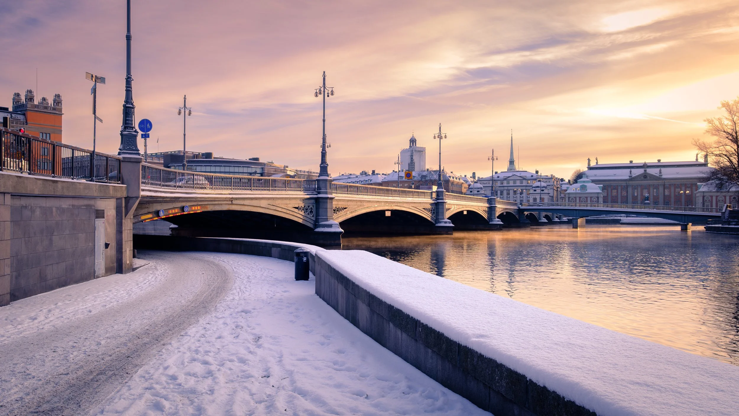 Ponte Vasabron em Estocolmo ao pôr do sol no inverno, com neve junto ao canal e edifícios históricos ao fundo.
