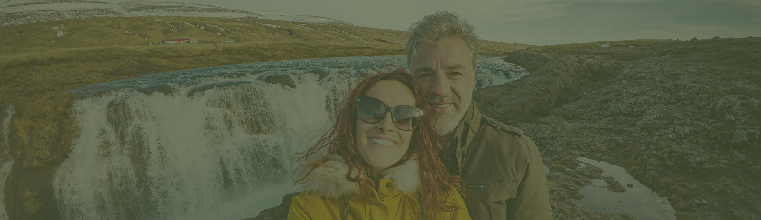 Casal em frente a uma cascata na Islândia, rodeado por uma paisagem rochosa e montanhosa.