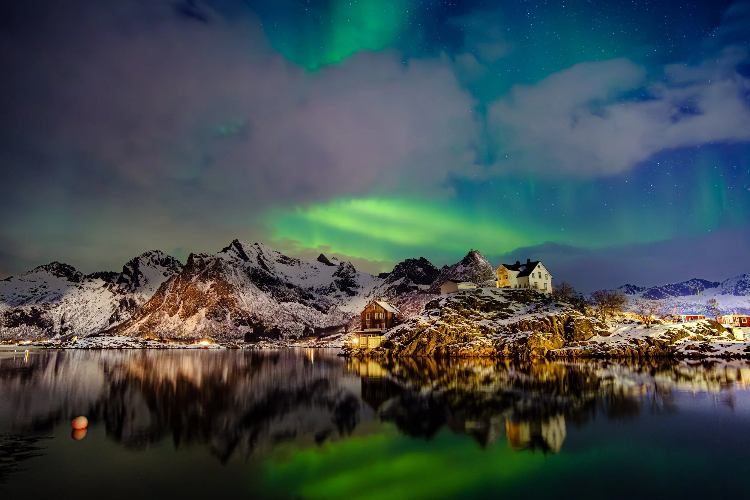 Casa de madeira junto a um fiorde norueguês, rodeada por montanhas nevadas refletidas na água.