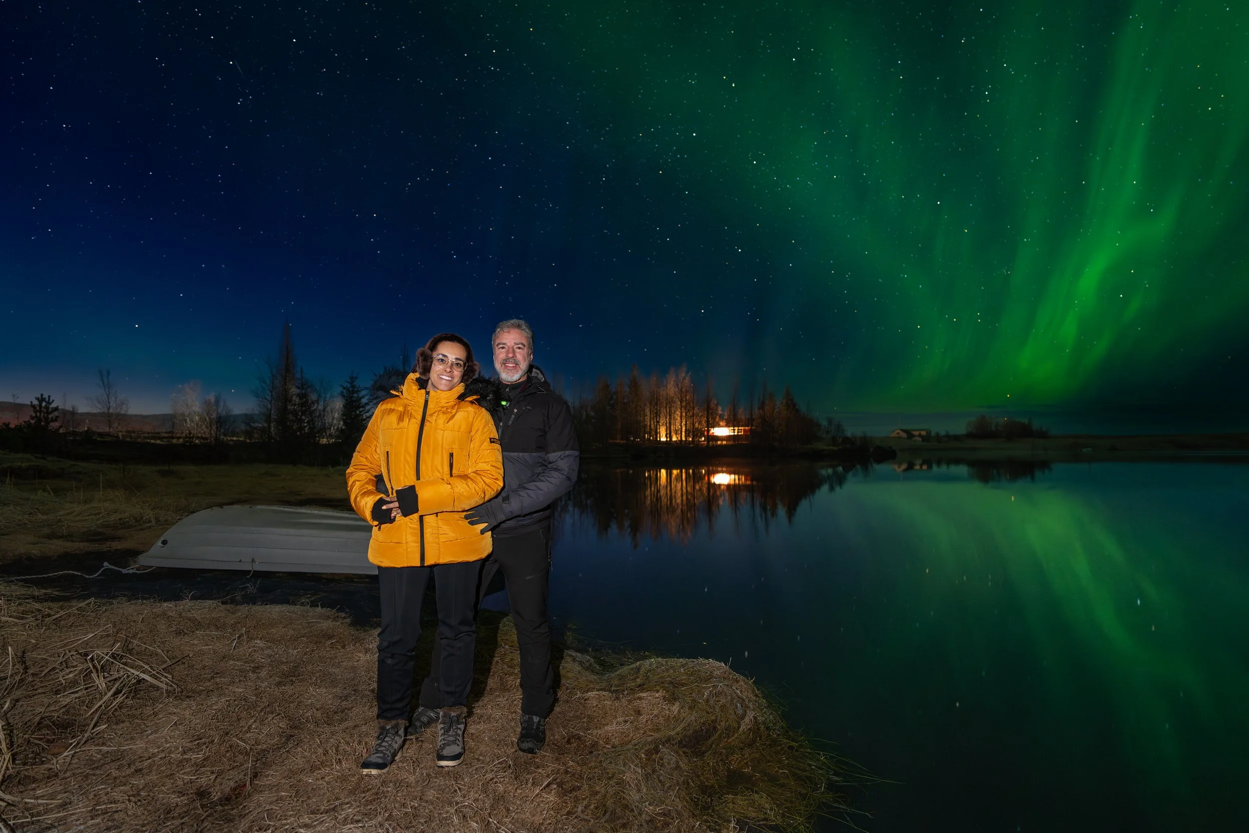 Casal junto a um lago sob a aurora boreal, com o reflexo das luzes verdes na água durante uma noite nórdica.
