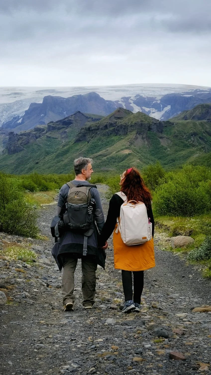 Casal a caminhar de mãos dadas por um trilho com montanhas e um glaciar na Islândia.