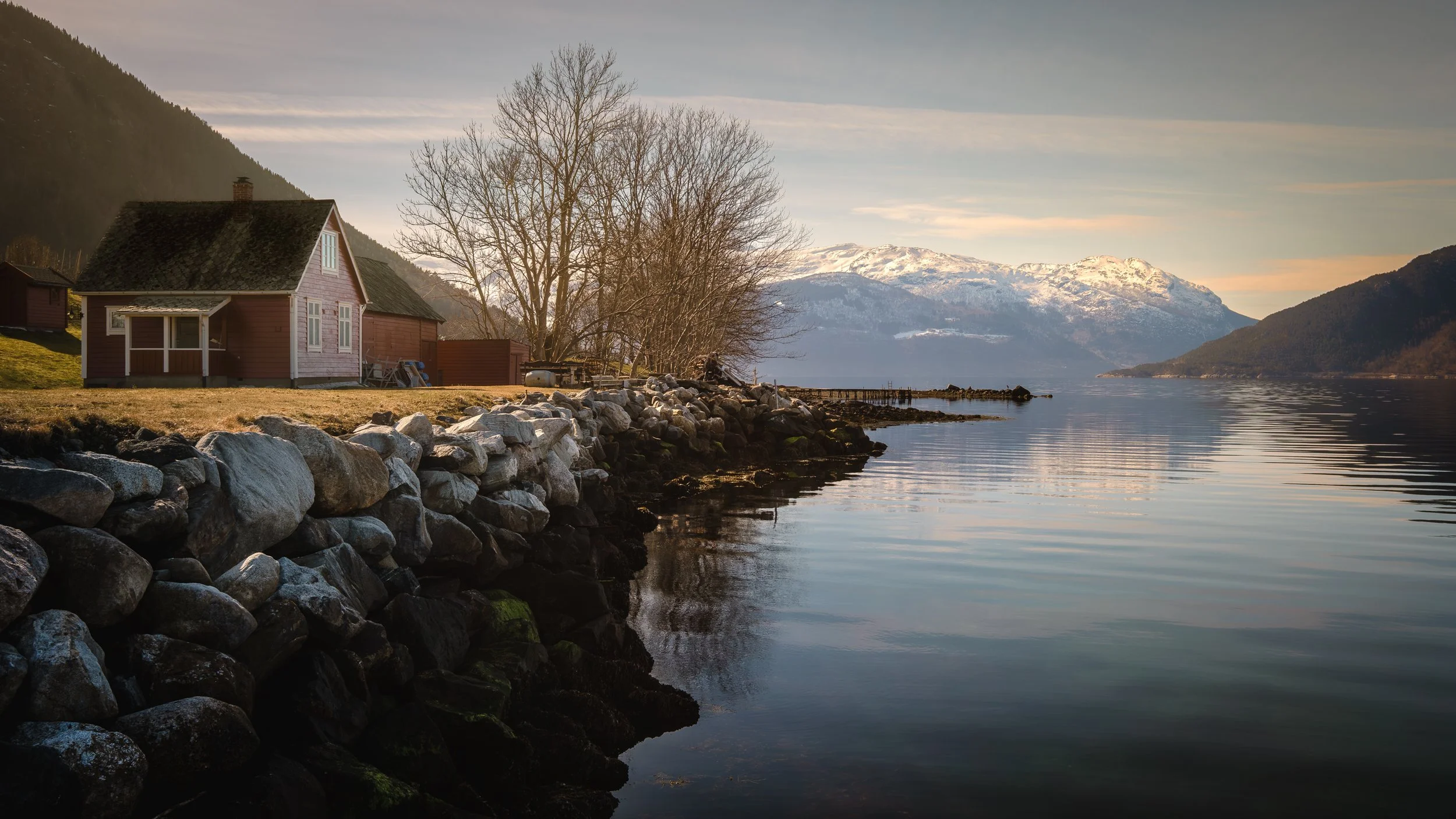 Casa de madeira junto a um fiorde norueguês, rodeada por montanhas nevadas refletidas na água.