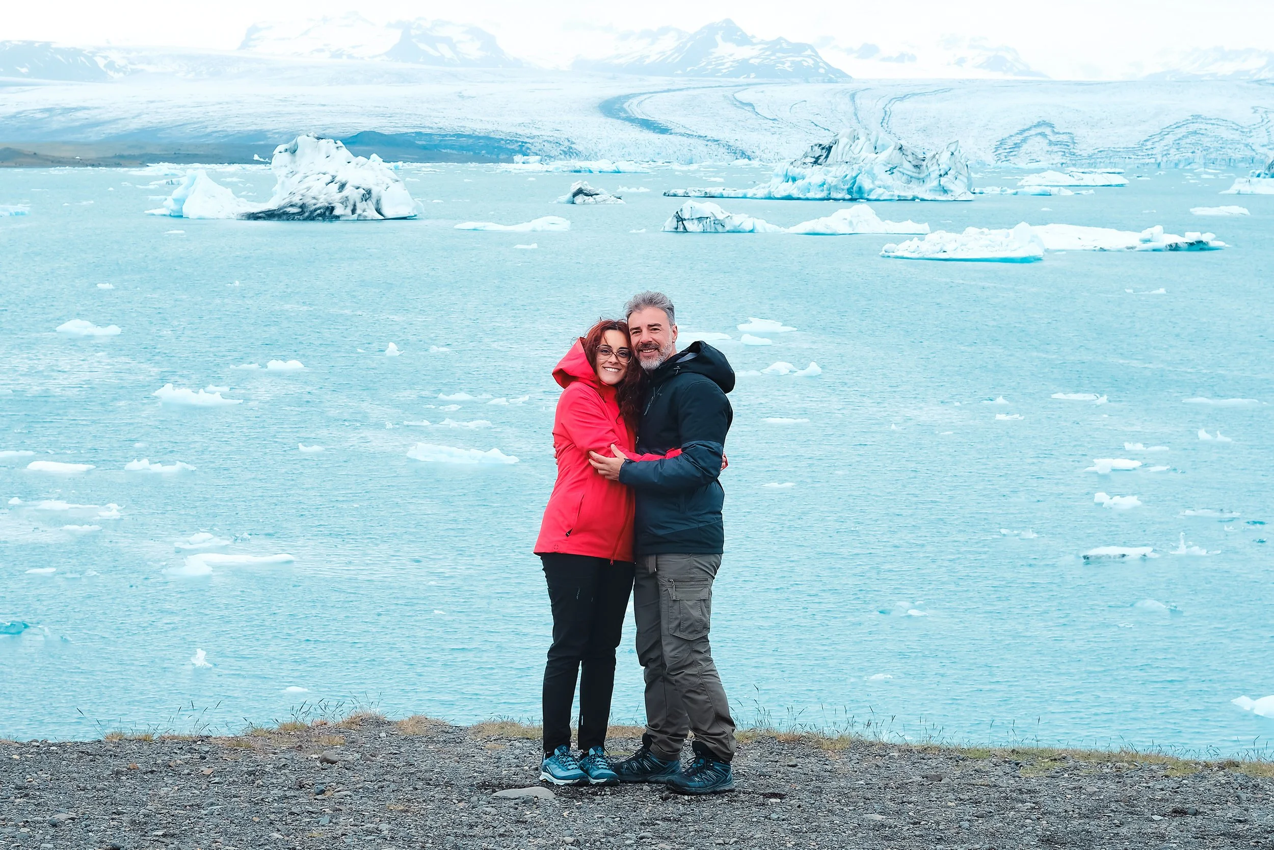 Casal abraçado junto à lagoa glaciar Jökulsárlón, na Islândia, com icebergs e o glaciar ao fundo.