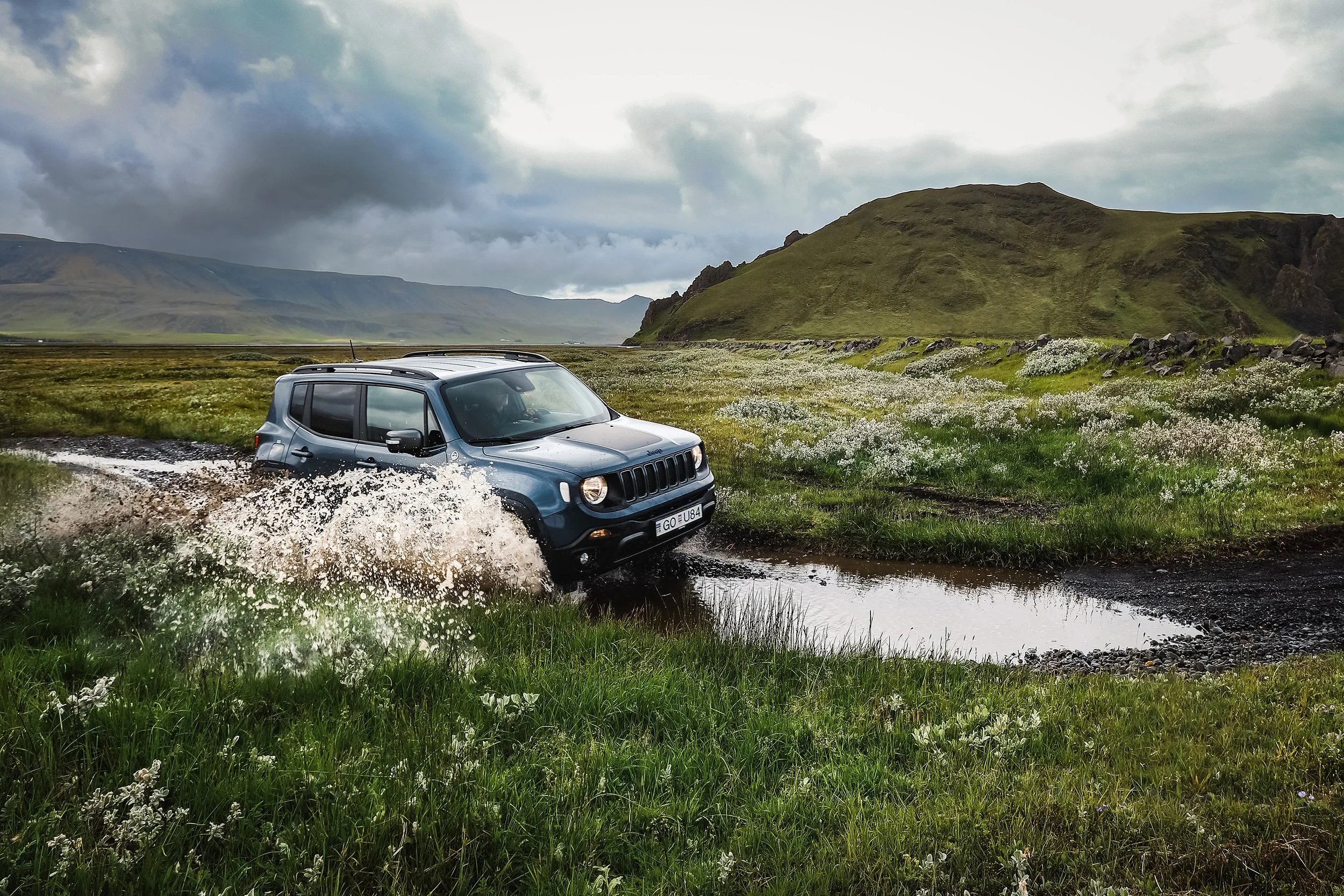 Carro 4x4 a atravessar um caminho com água numa paisagem verde e montanhosa da Islândia.