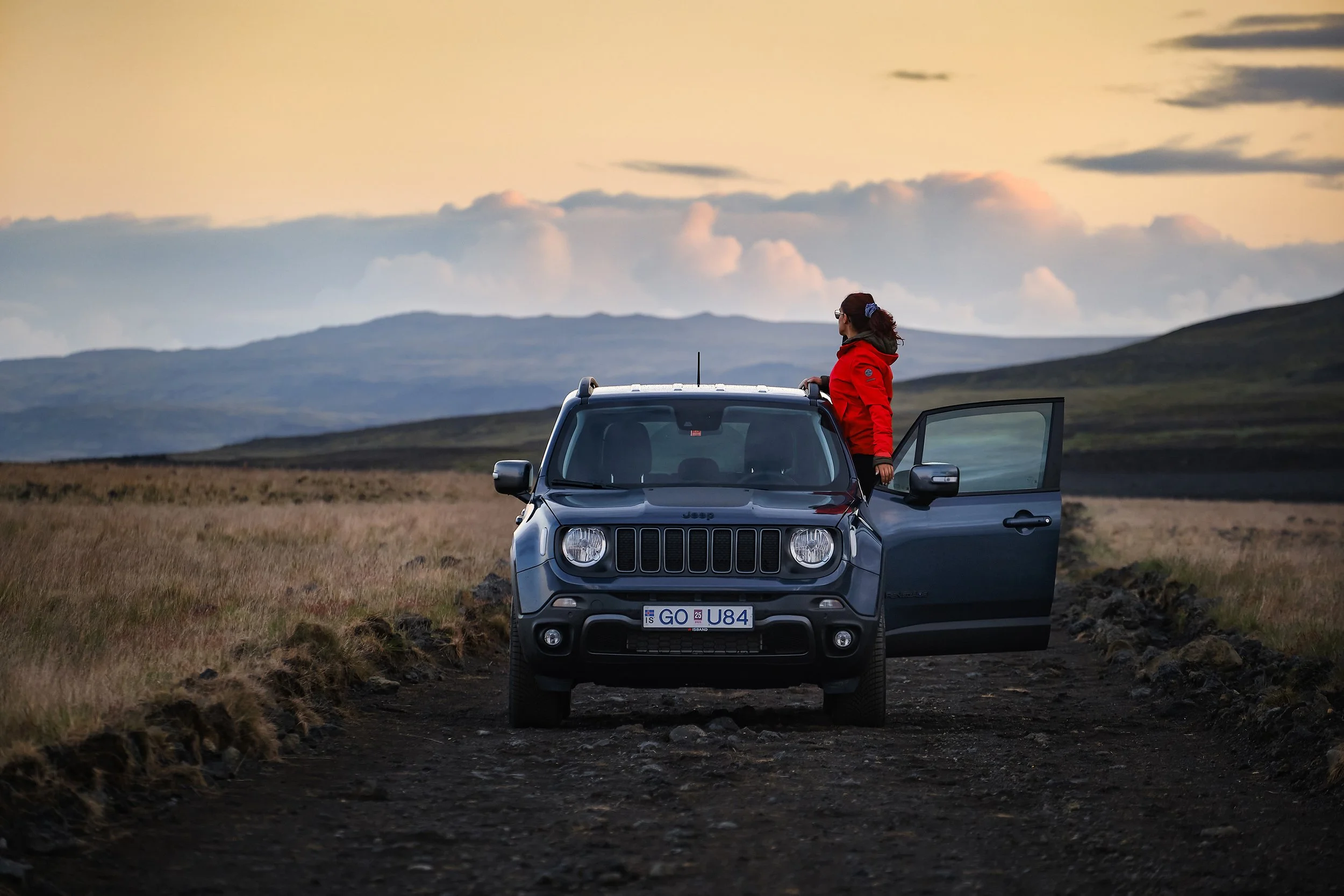 Viajante junto a um carro 4x4 numa estrada de terra, com montanhas ao fundo na Islândia ao pôr do sol.