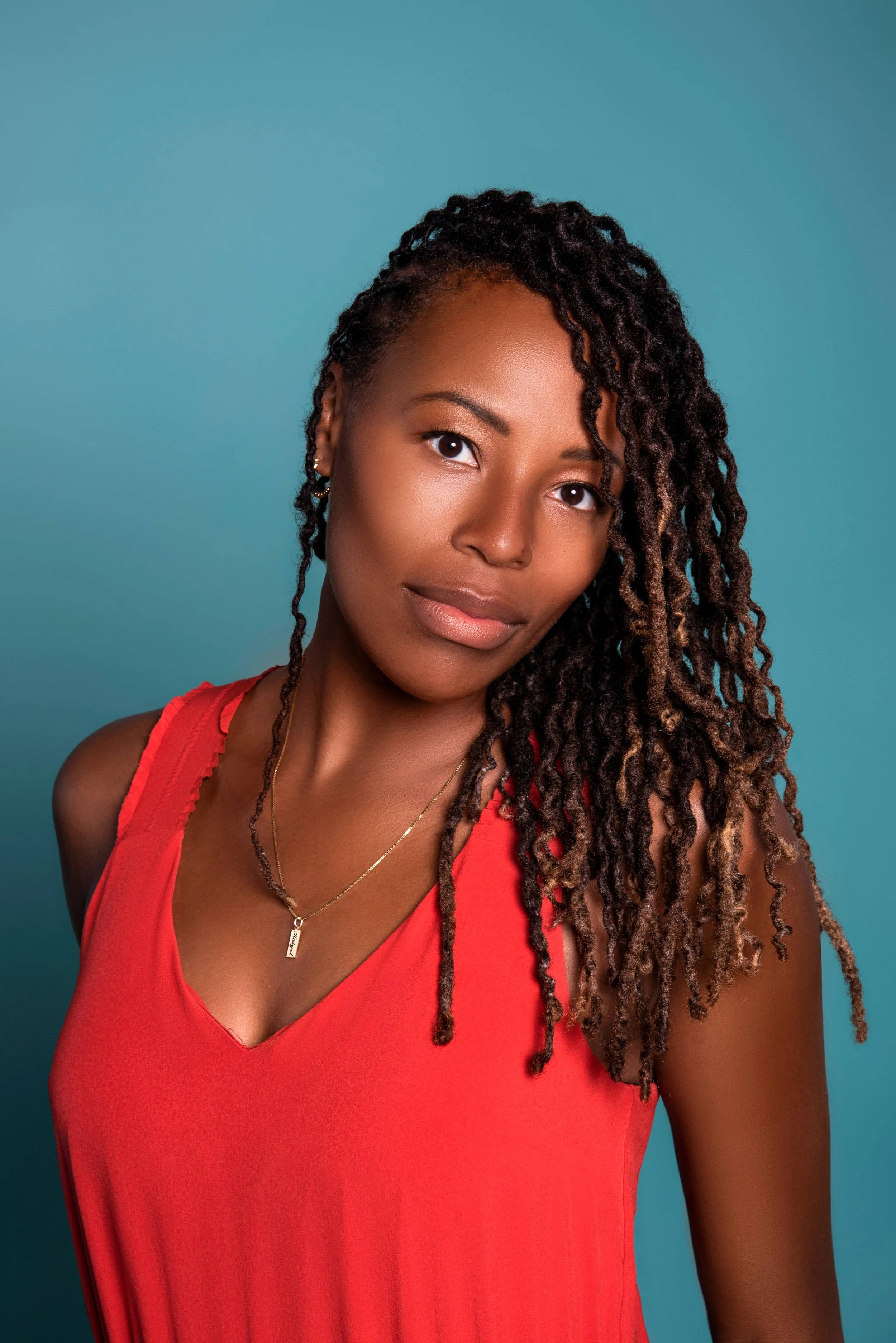 A woman with dark curly hair wearing a sleeveless red top and a gold necklace against a teal background.