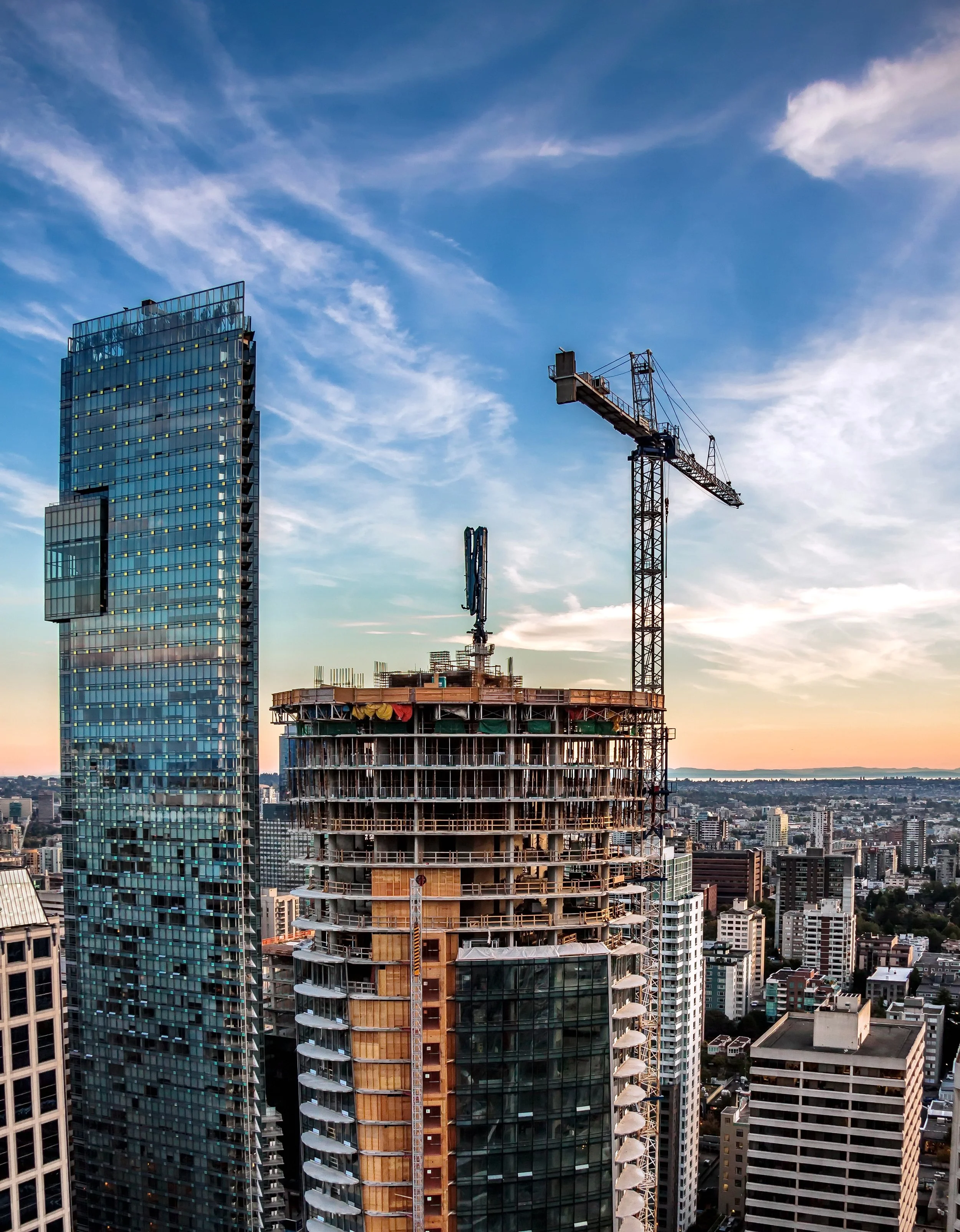 High-rise city buildings under a blue sky with wispy clouds, including a modern glass skyscraper and a building under construction with cranes.