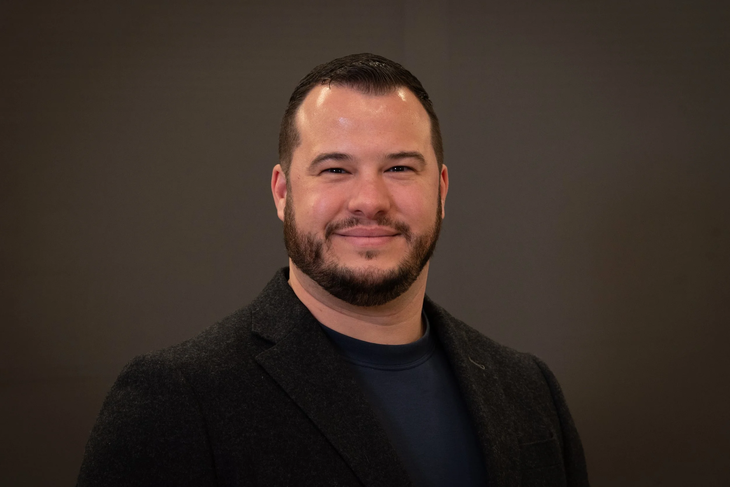 A man with dark hair, a beard, and a mustache, wearing a dark blazer and a black shirt, smiling against a gray background.