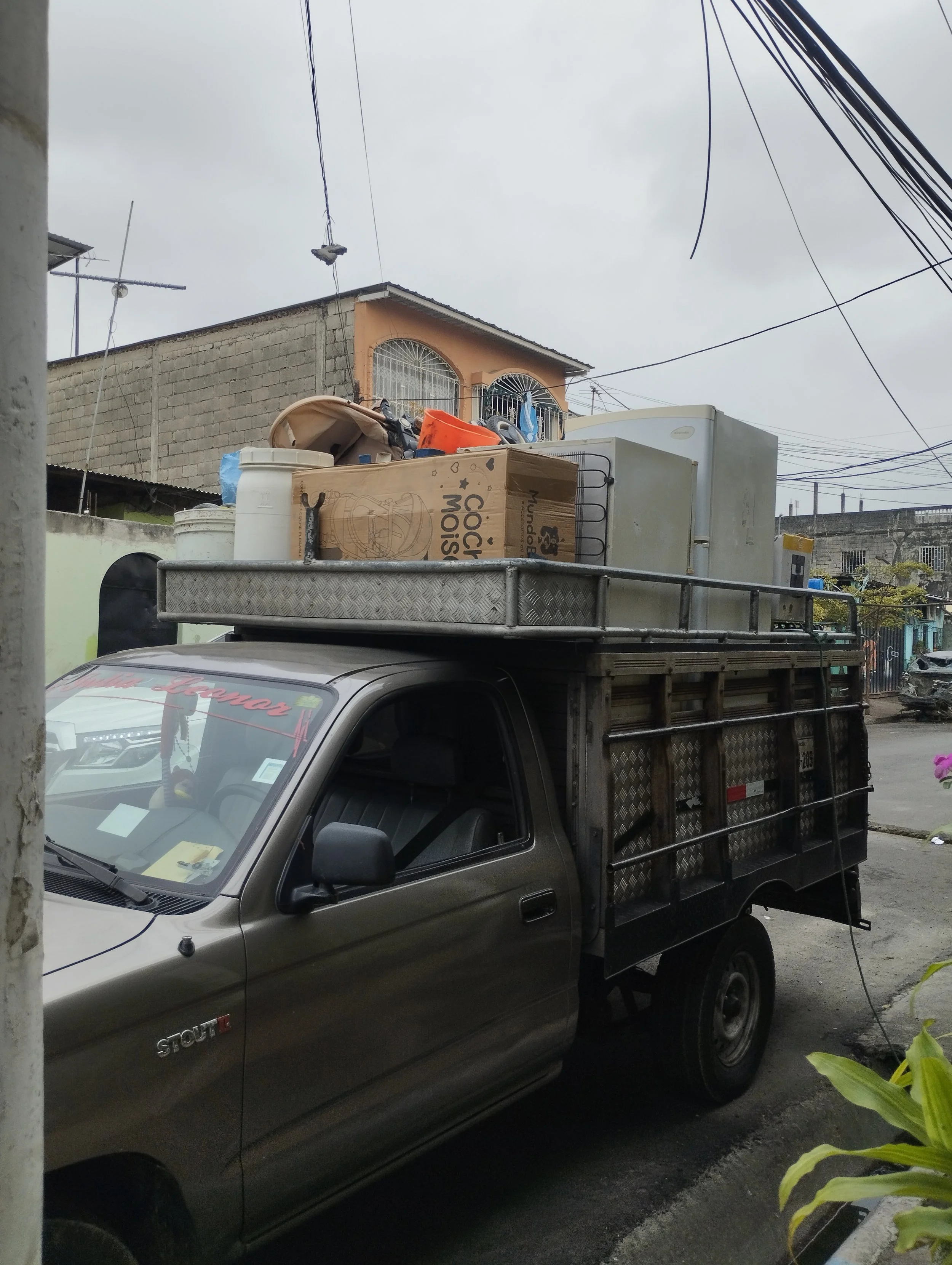 Camioneta de color gris con carga en la parte superior, en un barrio con edificios de fondo y cables eléctricos en el cielo nublado.