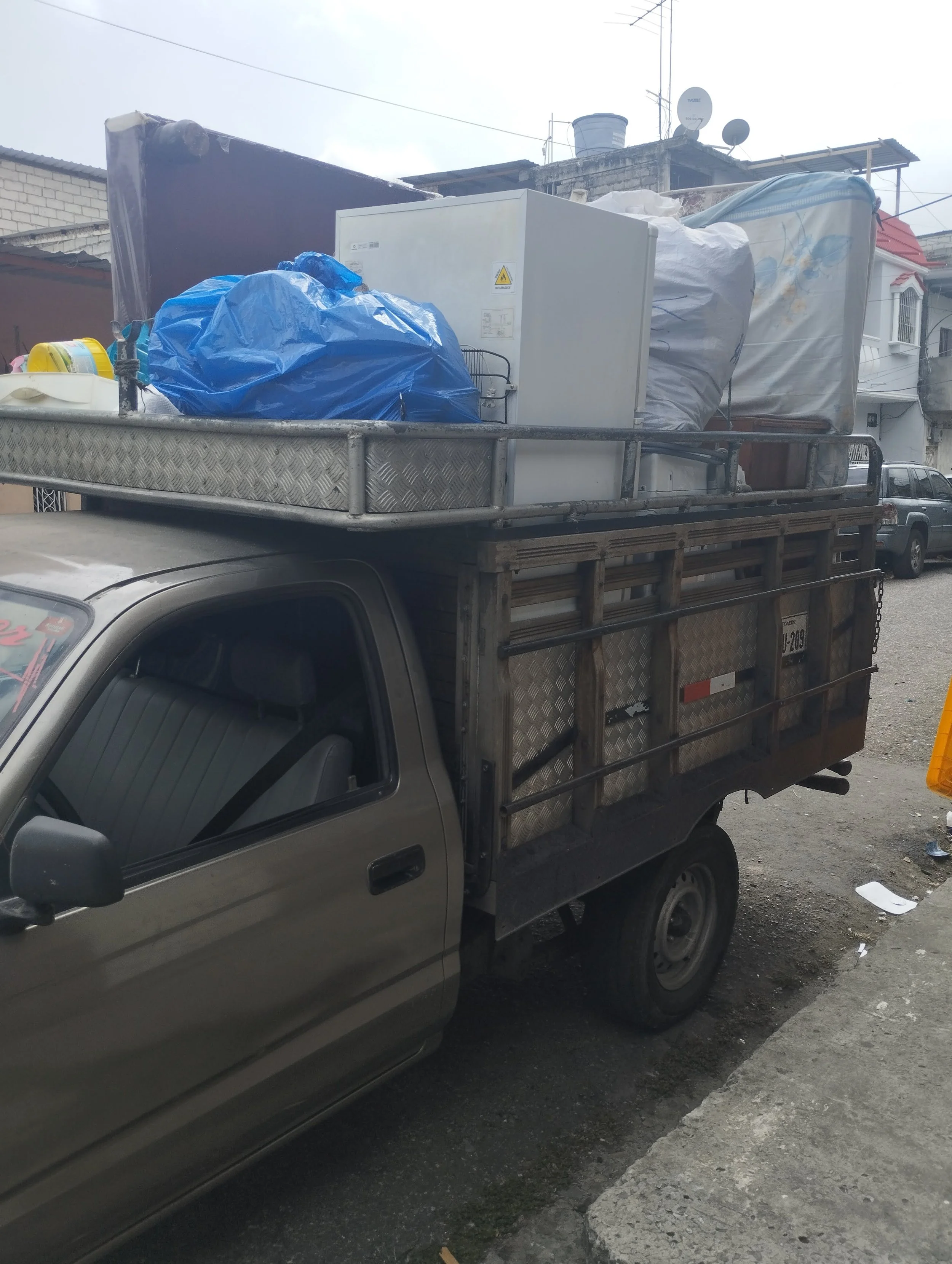 Camión pequeño de color beige cargado con varios objetos, incluyendo una nevera, un colchón y bolsas de basura, estacionado en la calle en un vecindario urbano.
